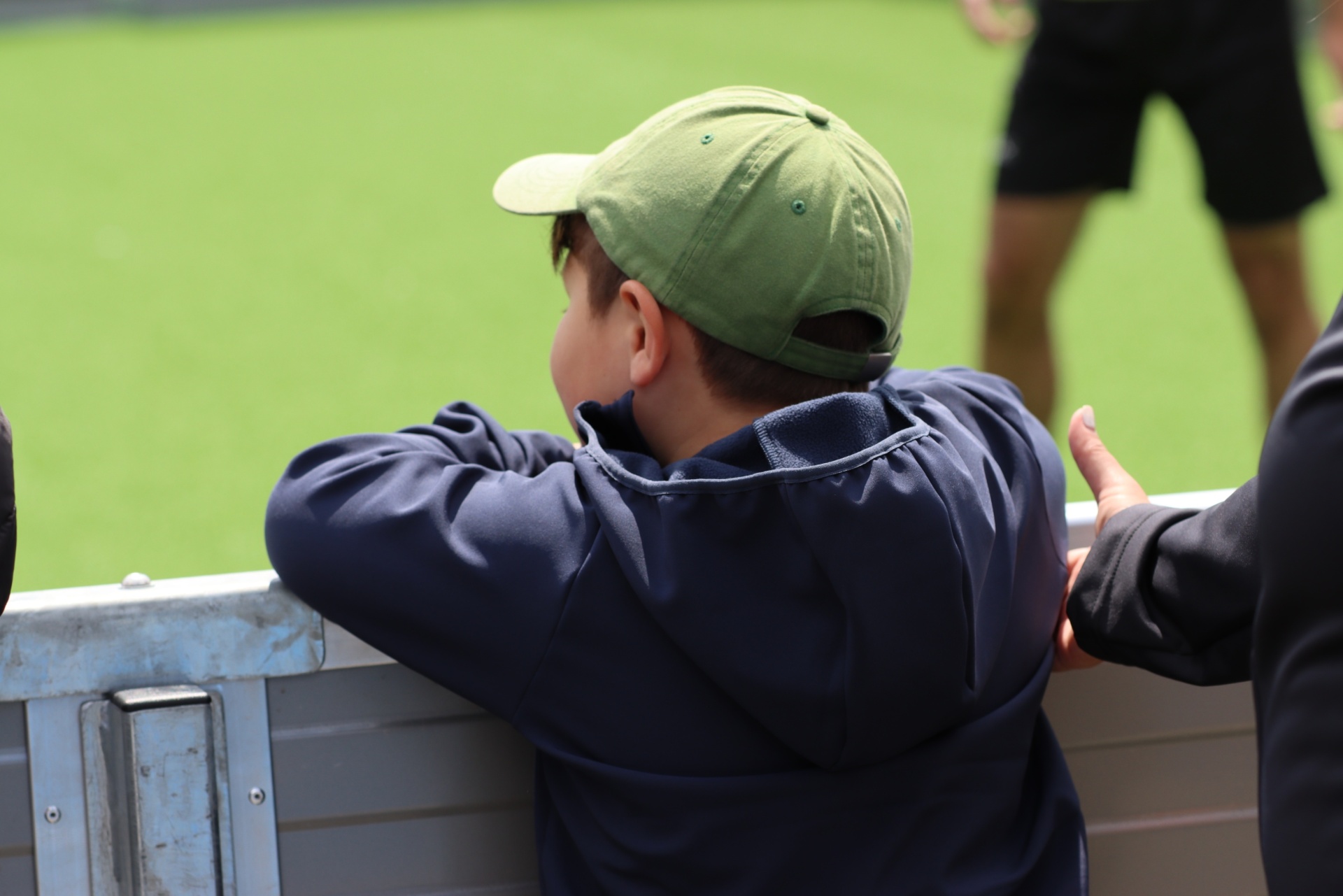 [{"clang_id":1,"value":"Junge mit Kappe sitzt am Rand einer Sportfläche, Blickrichtung nach vorne. Neben ihm eine weitere Person. Grüner Rasen im Hintergrund."}]