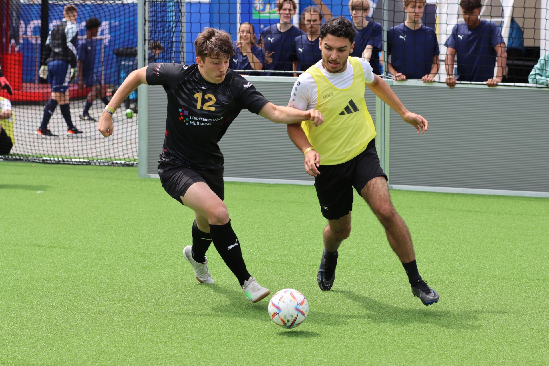 [{"clang_id":1,"value":"Zwei Fussballspieler auf grünem Kunstrasen im Wettkampf. Links Spieler mit Nummer 12 in schwarzem Trikot, rechts in gelbem Trikot und Weste. Im Hintergrund Zuschauer."}]
