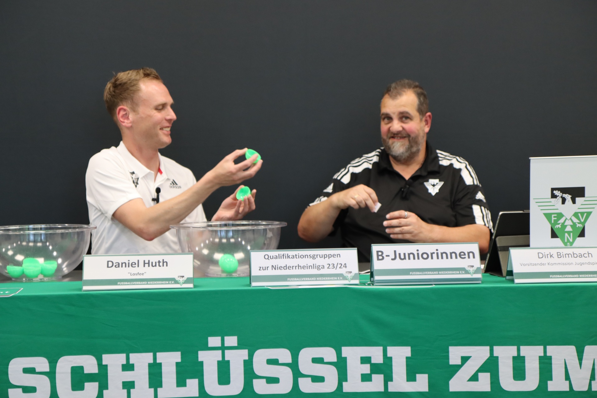 [{"clang_id":1,"value":"Zwei Männer sitzen an einem Tisch mit Namensschildern und ziehen Bälle aus transparenten Behältern. Im Hintergrund ein Banner mit dem Text \"SCHLÜSSEL ZUM\"."}]