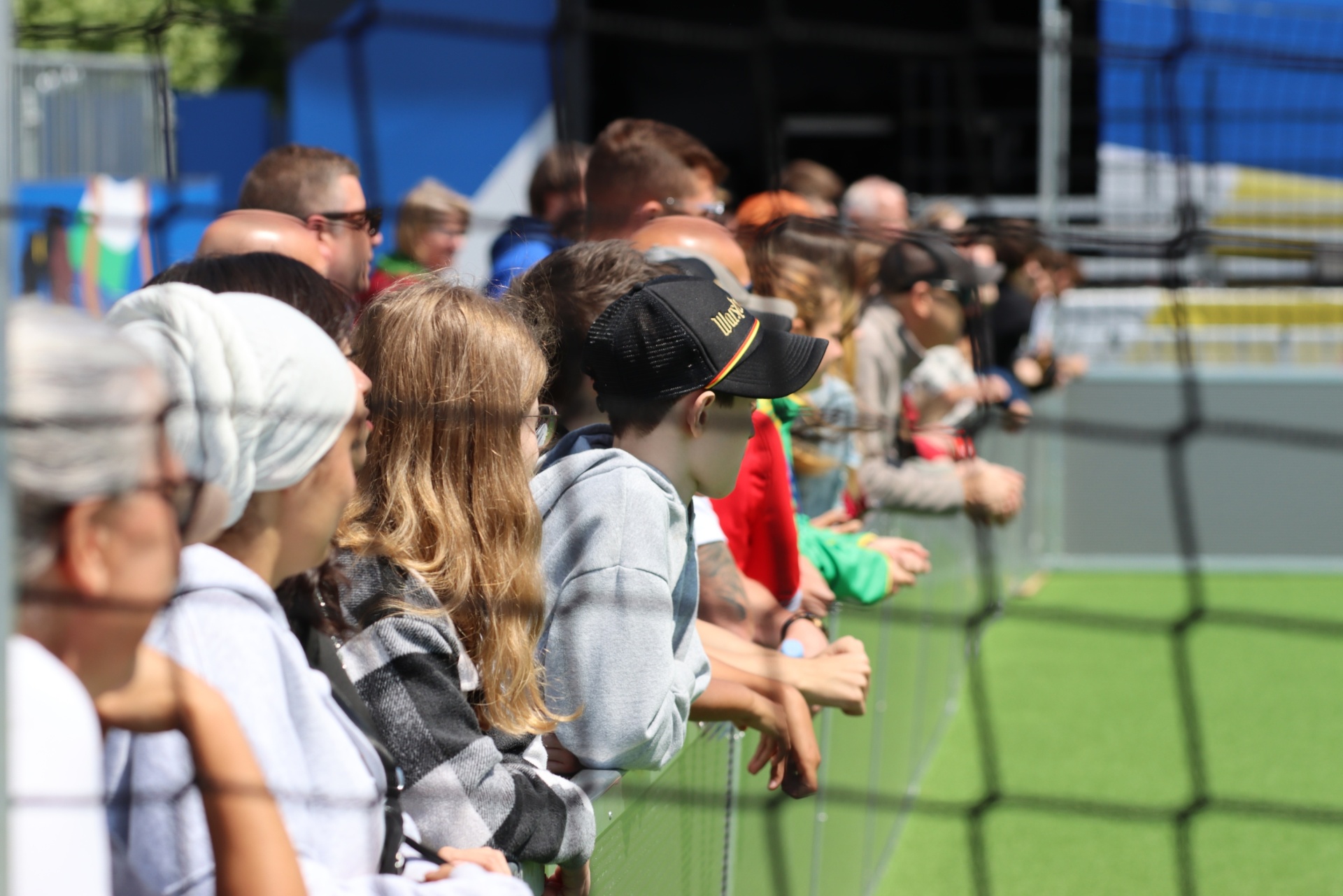 [{"clang_id":1,"value":"Zuschauerreihe vor einem Sportfeld. Kinder und Erwachsene sitzen oder stehen an einem Netz und schauen zu. Sonnenschein, blaues Hintergrundelement."}]