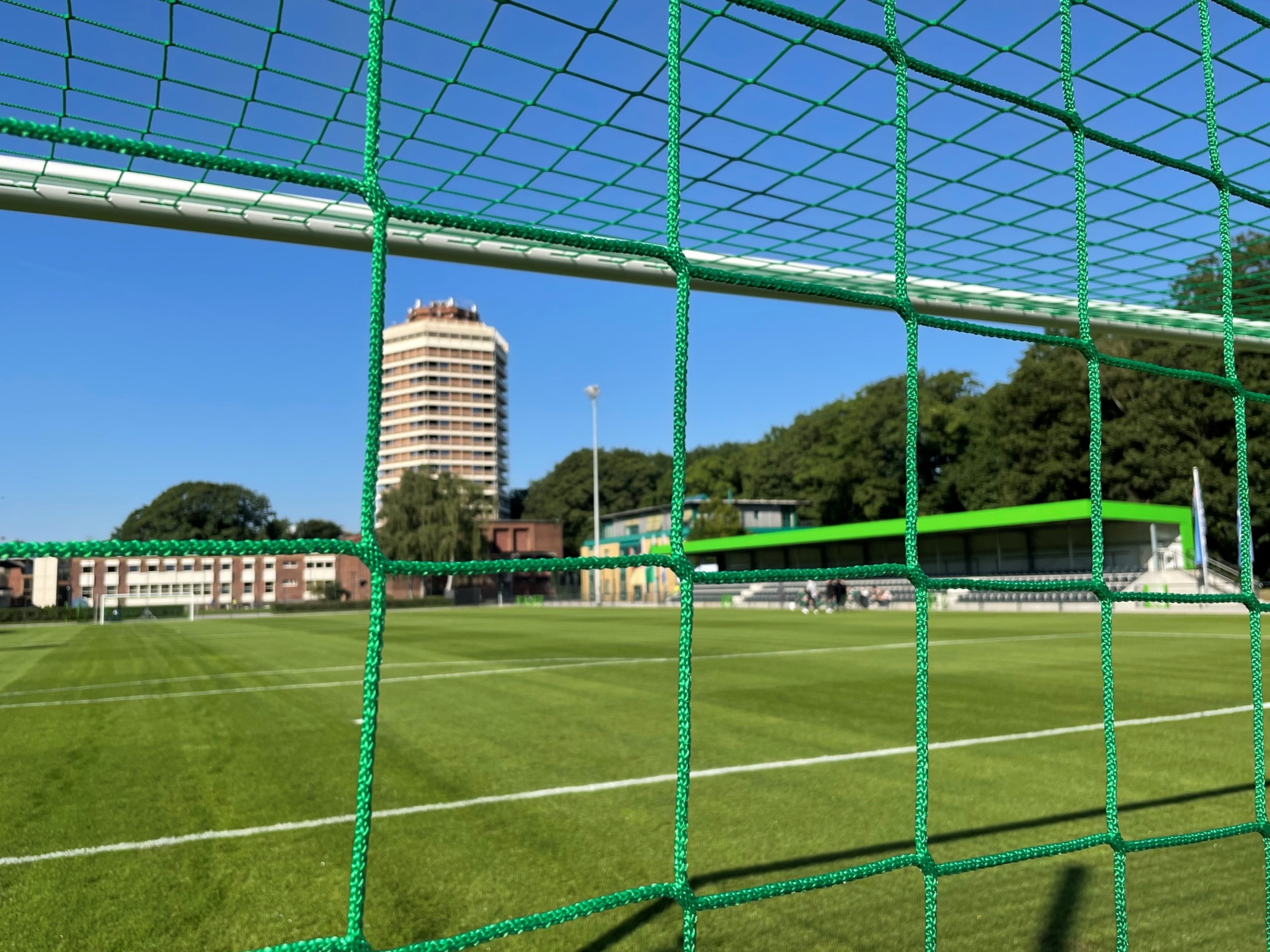 [{"clang_id":1,"value":"Fußballtor mit Netz im Fokus. Dahinter grüner Rasen, Stadiontribüne und hoher Wohnturm unter blauem Himmel."}]
