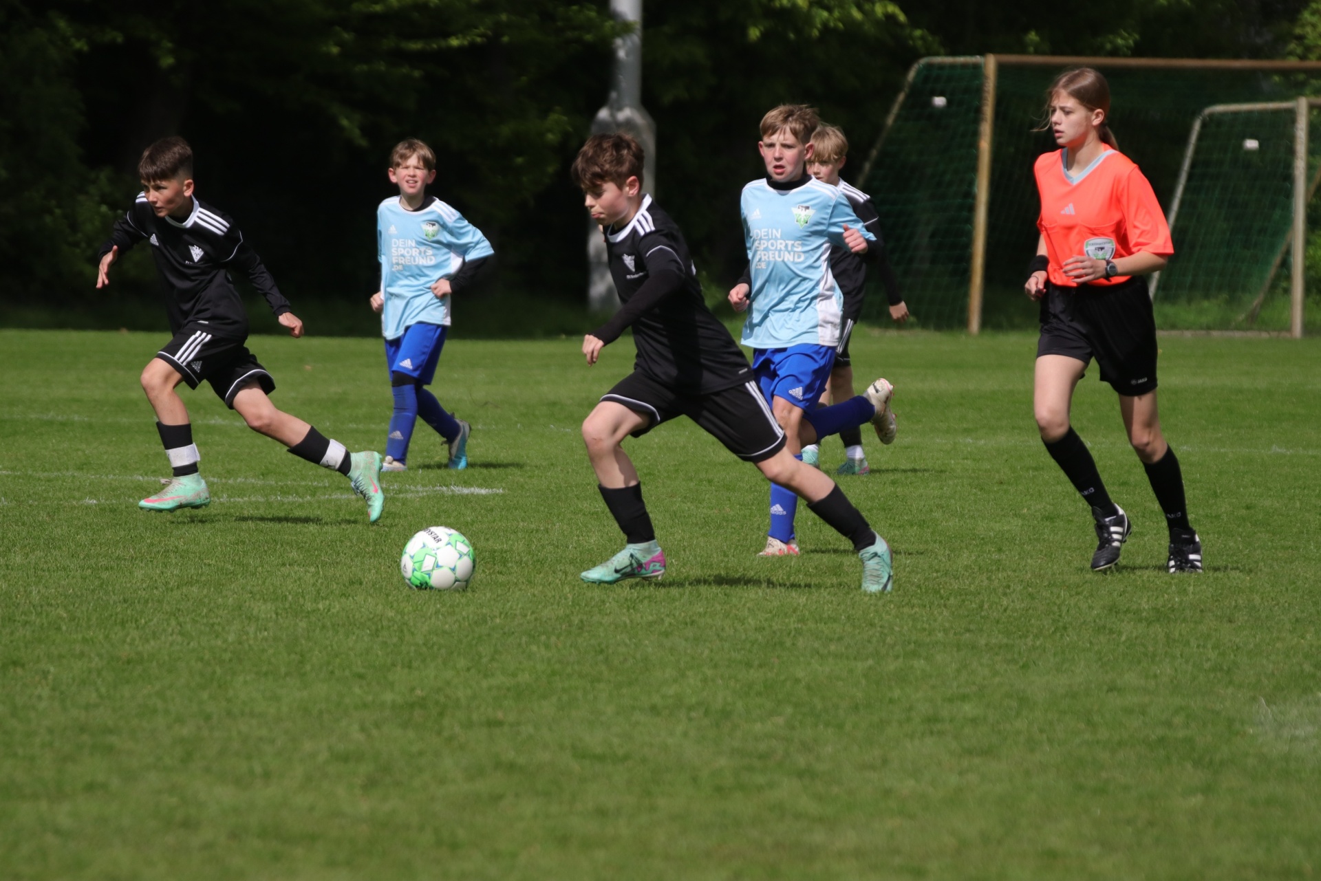 [{"clang_id":1,"value":"Jugendfußballspiel auf Grasplatz. Mehrere Spieler in hellblauen und schwarz-weißen Trikots verfolgen den Ball. Rechts eine Schiedsrichterin in orange."}]