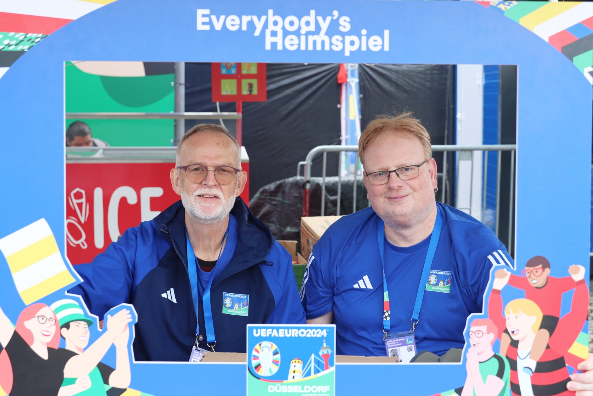 [{"clang_id":1,"value":"Zwei Männer vor blauem Werbeaufsteller mit Schriftzug \"Everybody's Heimspiel\" und UEFA EURO 2024 Logo. Beide tragen blaue Sportjacken und Lanyards. Im Hintergrund Menschen und Stadionelemente."}]