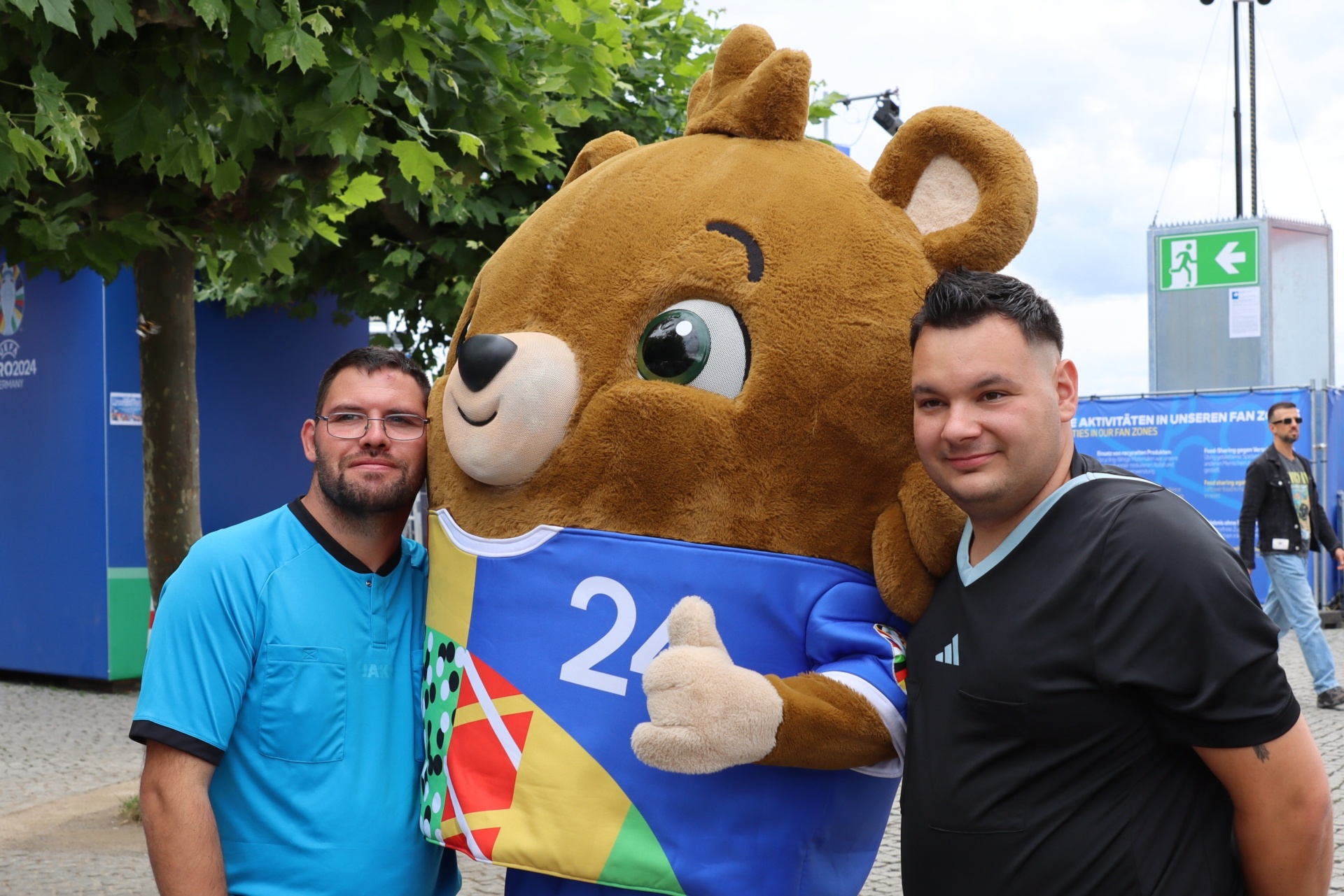 [{"clang_id":1,"value":"Zwei Männer stehen mit einem braunen Maskottchen-Bären mit blauem Trikot (Zahl 24) vor einem Hintergrund mit EURO 2024-Werbung."}]