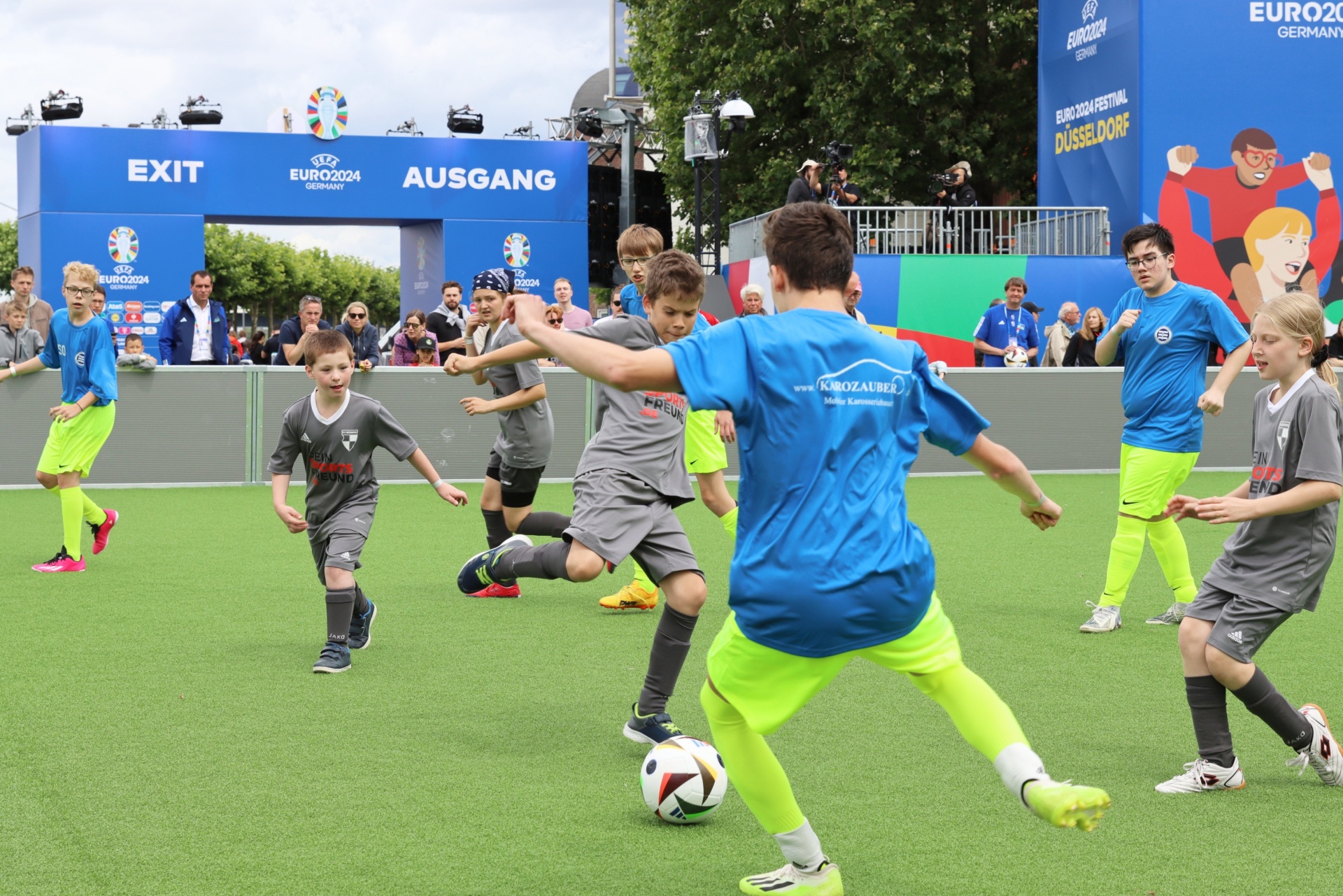[{"clang_id":1,"value":"Fußballspiel mit Kindern auf grünem Kunstrasen. Im Hintergrund eine Stadionkulisse mit EURO2024-Werbung und Menschen."}]