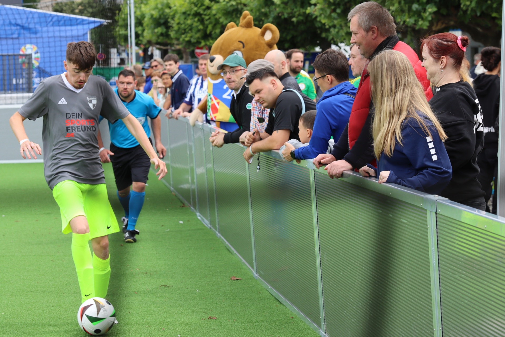 [{"clang_id":1,"value":"Fußballer jongliert Ball auf grünem Kunstrasen. Im Hintergrund Zuschauer hinter einer Absperrung, darunter ein Mann mit blauem Pullover und eine Frau mit blonden Haaren."}]