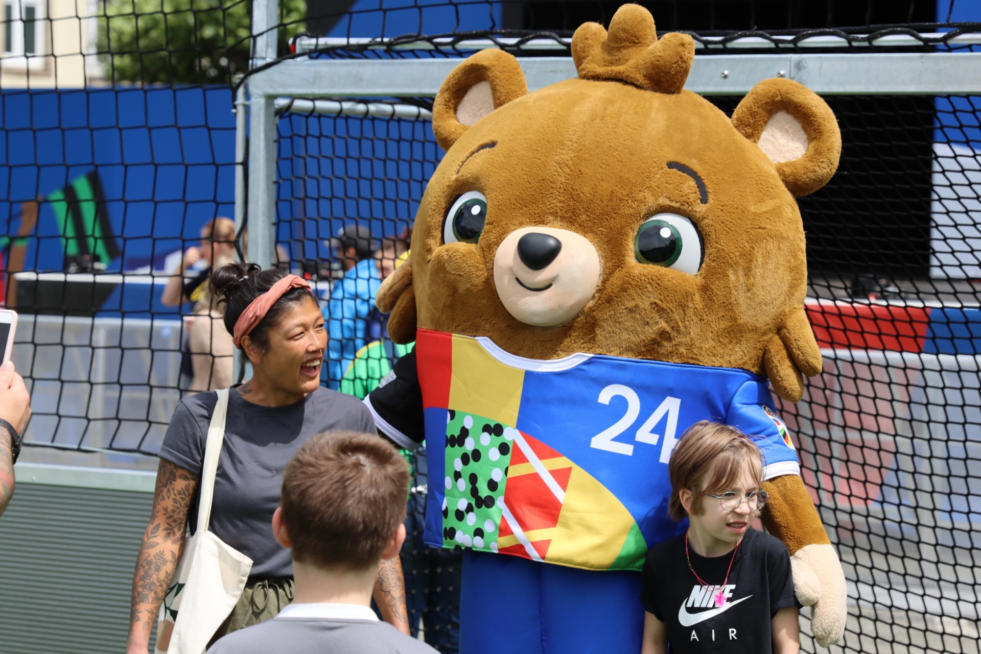 [{"clang_id":1,"value":"Brauner Bär in Sportkleidung mit Nummer 24 umarmt eine Frau. Im Hintergrund ein Fußballtor und Menschen."}]