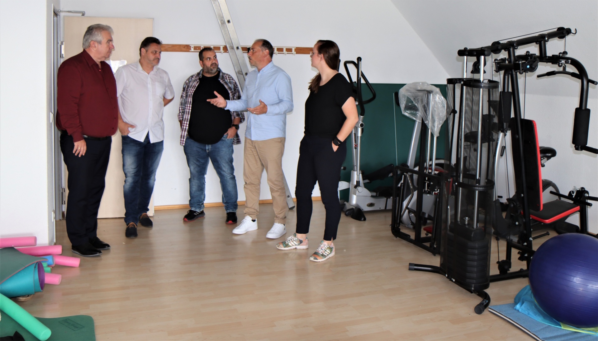 [{"clang_id":1,"value":"Fünf Personen stehen in einem hellen Raum mit Fitnessgeräten. Eine Frau erklärt den Männern etwas, während sie im Kreis diskutieren."}]
