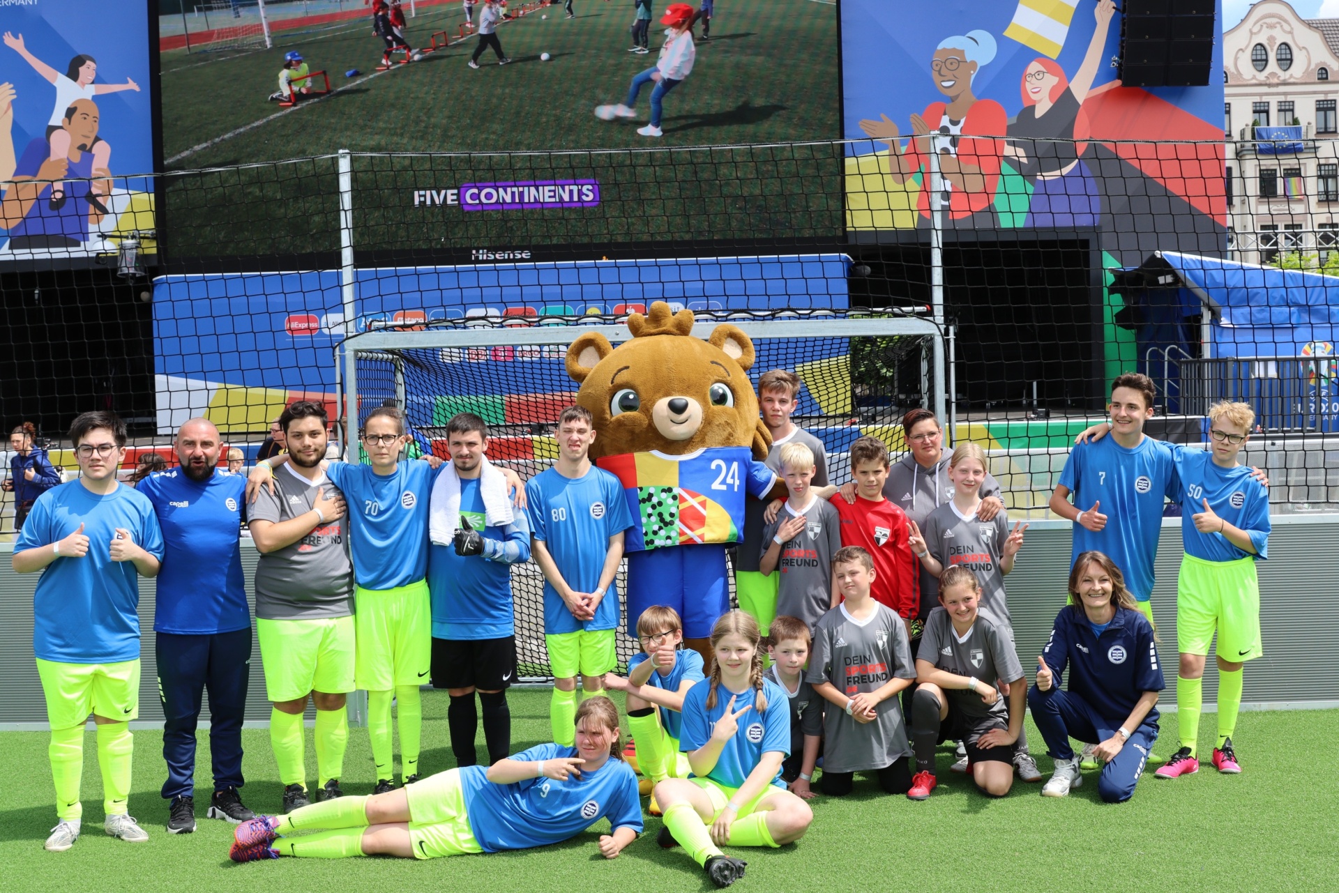 [{"clang_id":1,"value":"Fußballgruppe mit Kindern und Trainer vor einem Tor. Im Hintergrund eine Großbildleinwand mit Fußballspiel und Werbebanner. Ein Maskottchen ist ebenfalls zu sehen."}]