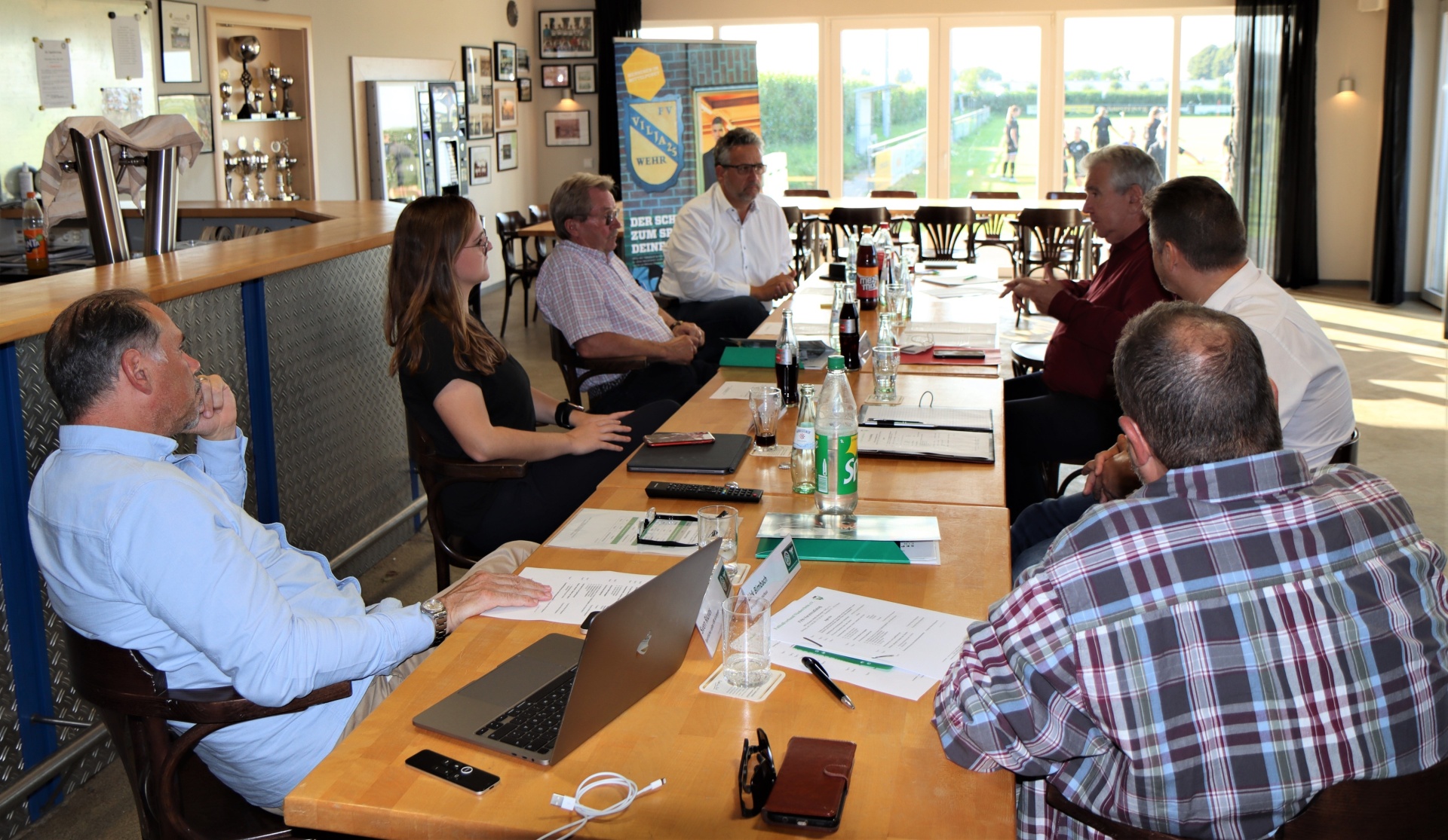 [{"clang_id":1,"value":"Gruppe diskutiert an einem Tisch in hellem Raum mit Blick auf Grünfläche. Vorhanden: Laptops, Notizen, Getränke. Im Hintergrund Bar und Trophäen."}]