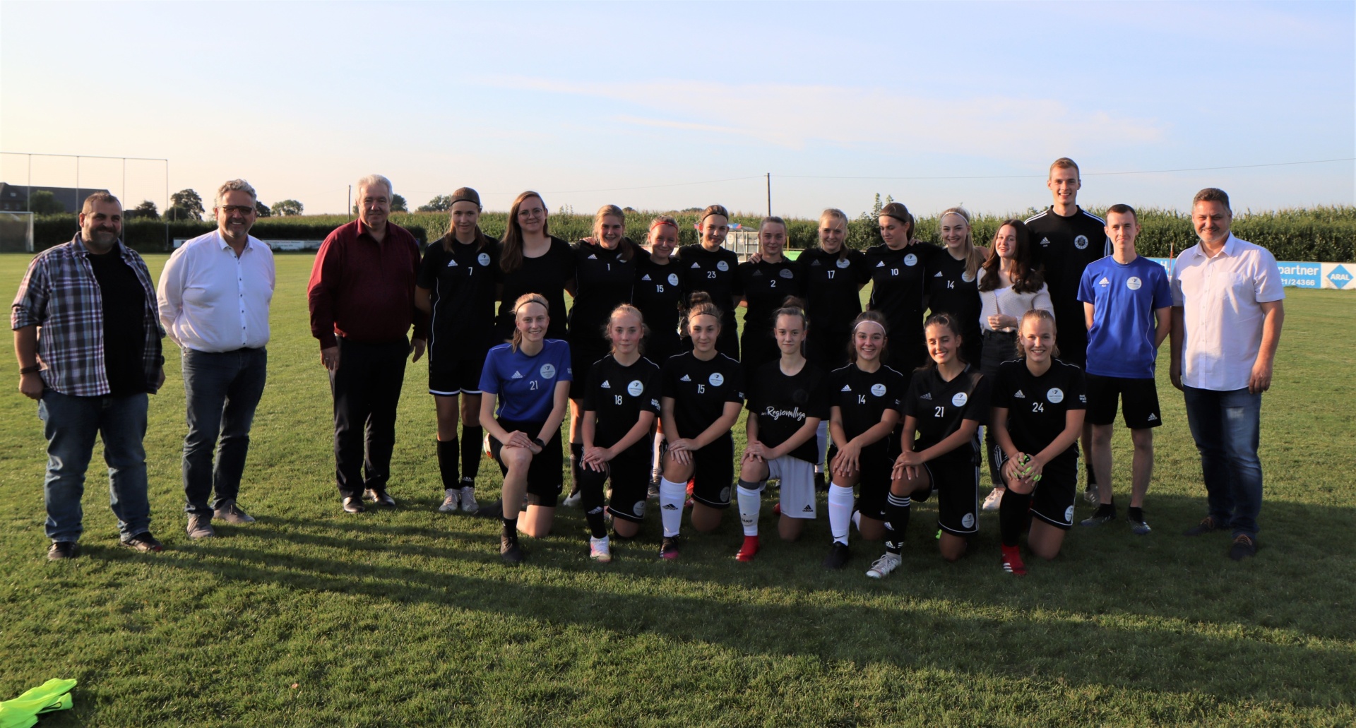 [{"clang_id":1,"value":"Fußballteam kniet und steht vor mehreren Männern und Frauen auf einem Rasenfeld. Im Hintergrund ein blauer Himmel und Maisfeld."}]