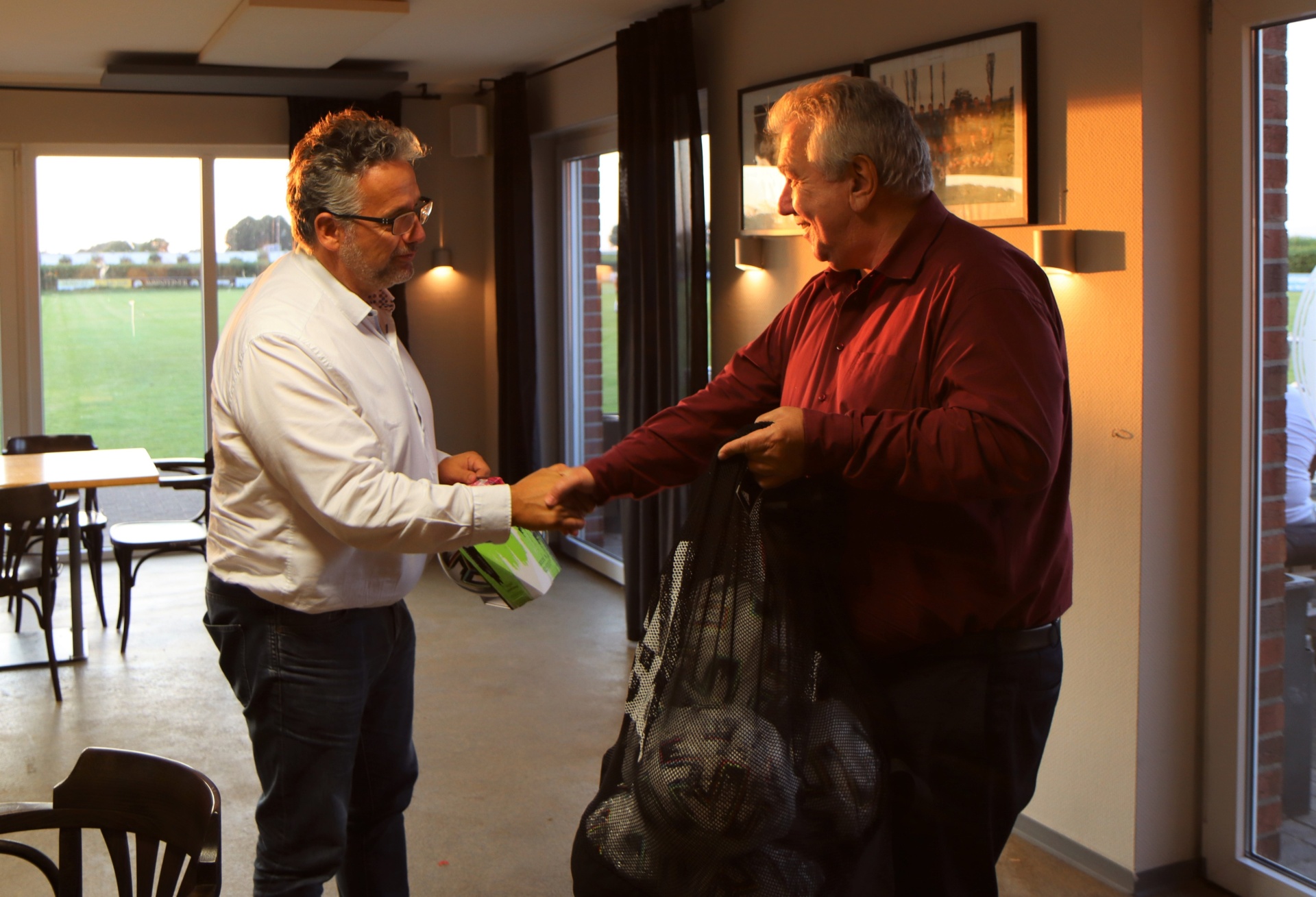 [{"clang_id":1,"value":"Zwei Männer im Innenraum eines Gebäudes, wahrscheinlich einem Clubhaus. Einer reicht dem anderen eine kleine Schachtel, der andere hält einen Sack. Im Hintergrund ein Fußballfeld."}]