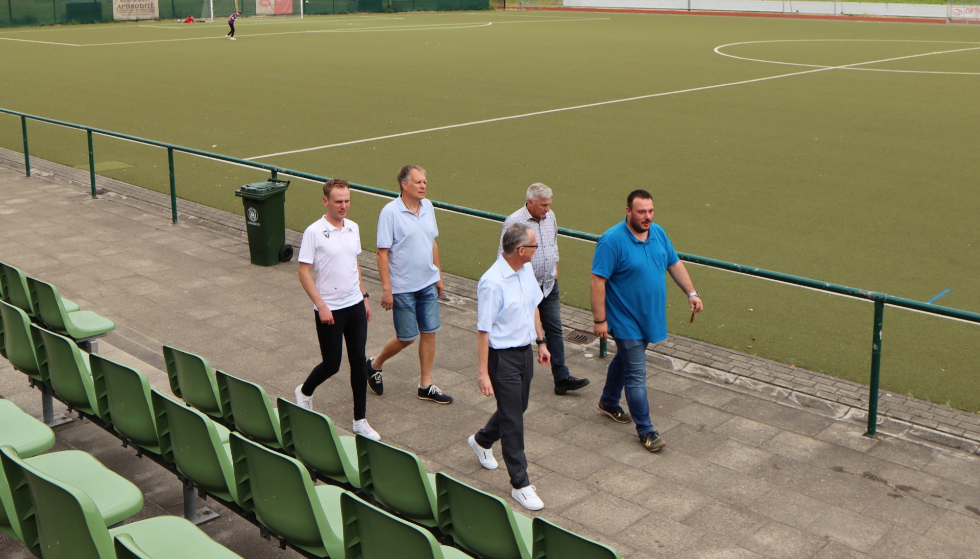 [{"clang_id":1,"value":"Vier Männer gehen an Zuschauerrängen eines Fußballfeldes vorbei. Das Spielfeld ist mit Kunstgras belegt."}]