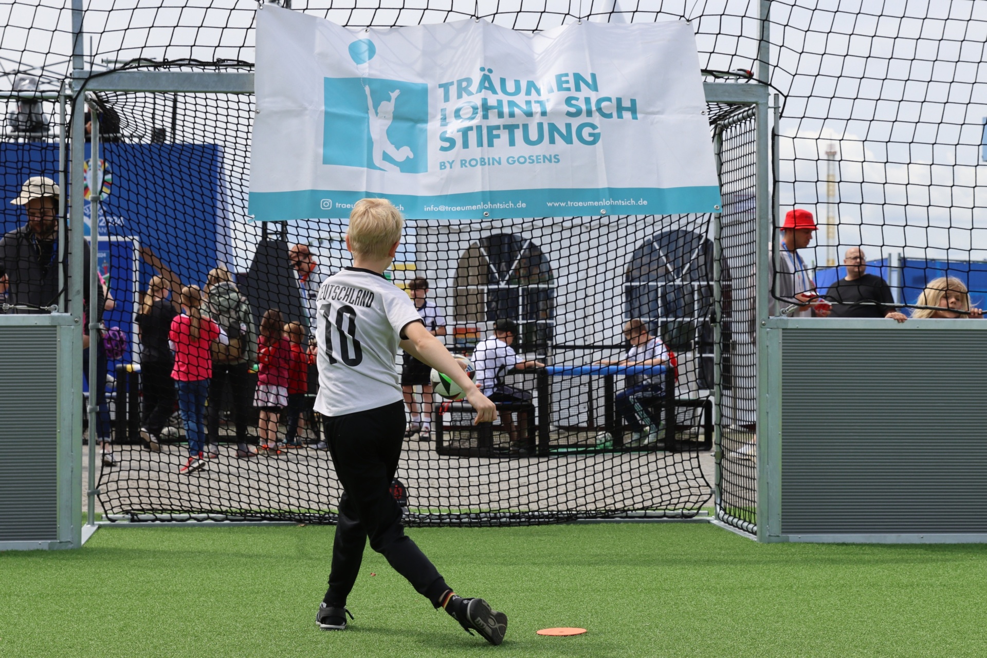 [{"clang_id":1,"value":"Fußballer in Deutschland-Trikot schießt auf Tor. Im Hintergrund ein Netz und Banner mit dem Aufdruck \"Träumen könnt sich Stiftung\". Menschen stehen zuschauend."}]