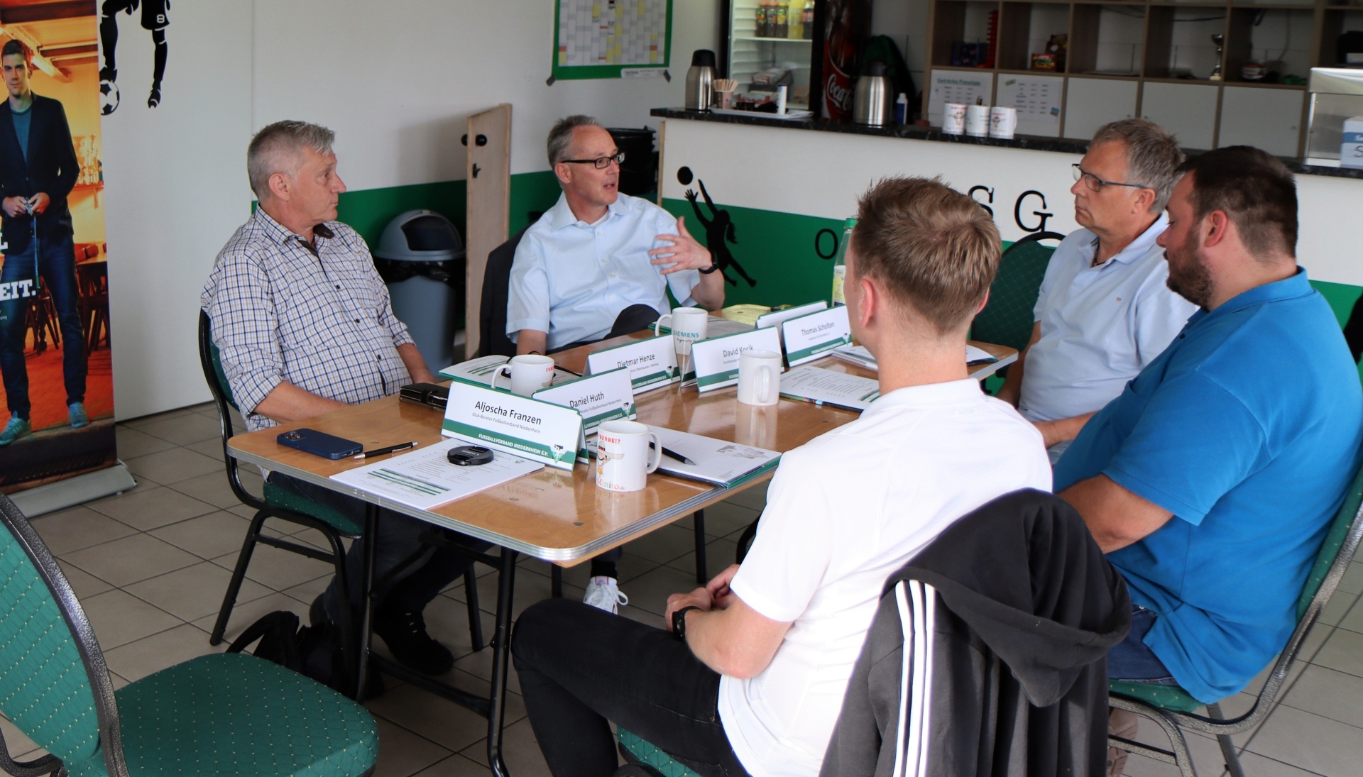 [{"clang_id":1,"value":"Fünf Männer sitzen an einem Konferenztisch in einem Raum mit Fliesenboden. Sie unterhalten sich, einige gestikulieren. Im Hintergrund eine Wand mit Getränken und ein Plakat."}]
