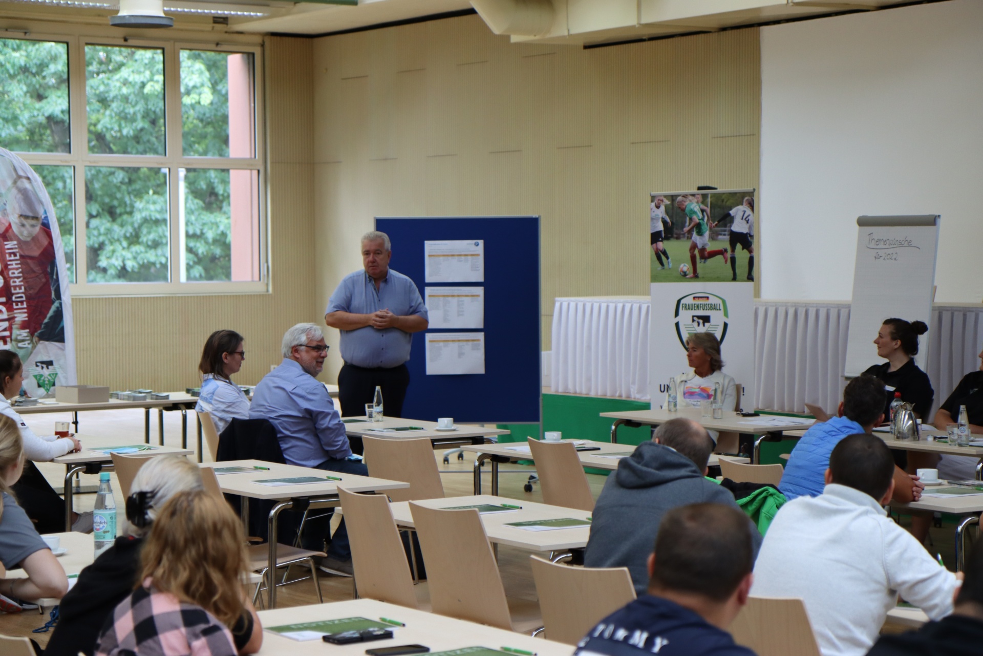 [{"clang_id":1,"value":"Ein Mann steht vor einer Gruppe sitzender Menschen in einem hellen Raum, wahrscheinlich eine Sporthalle oder ein Gemeinschaftszentrum. Tische mit Notizen und Stiften sind vor den Teilnehmern. Im Hintergrund hängen Poster, darunter eines mit Fußballspielern."}]