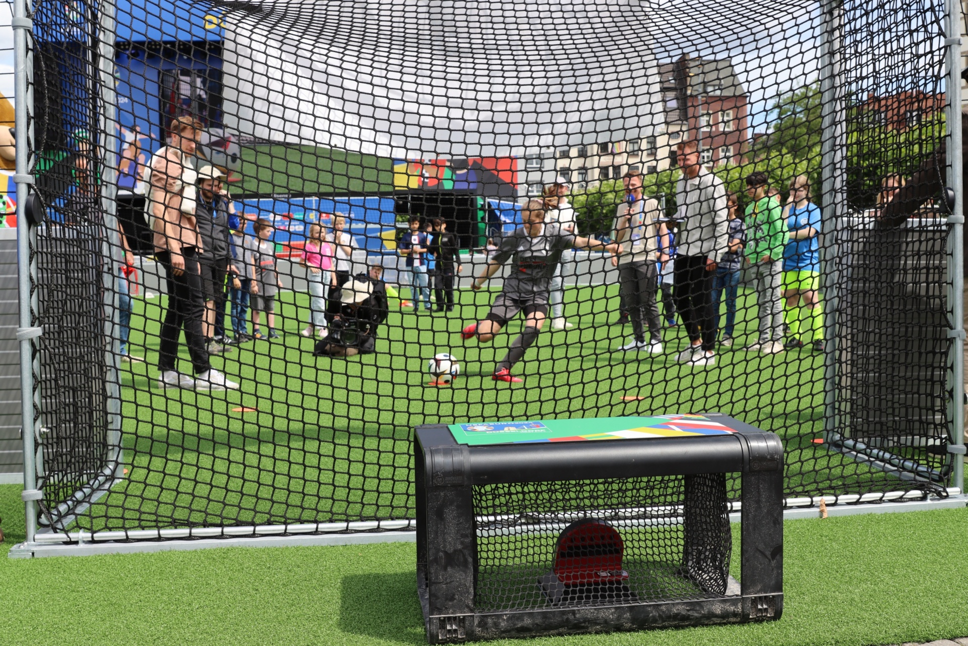 [{"clang_id":1,"value":"Fußball-Skillstation mit Netz und kleinem Tor im Vordergrund. Im Hintergrund spielende Kinder und Erwachsene auf grünem Rasen."}]