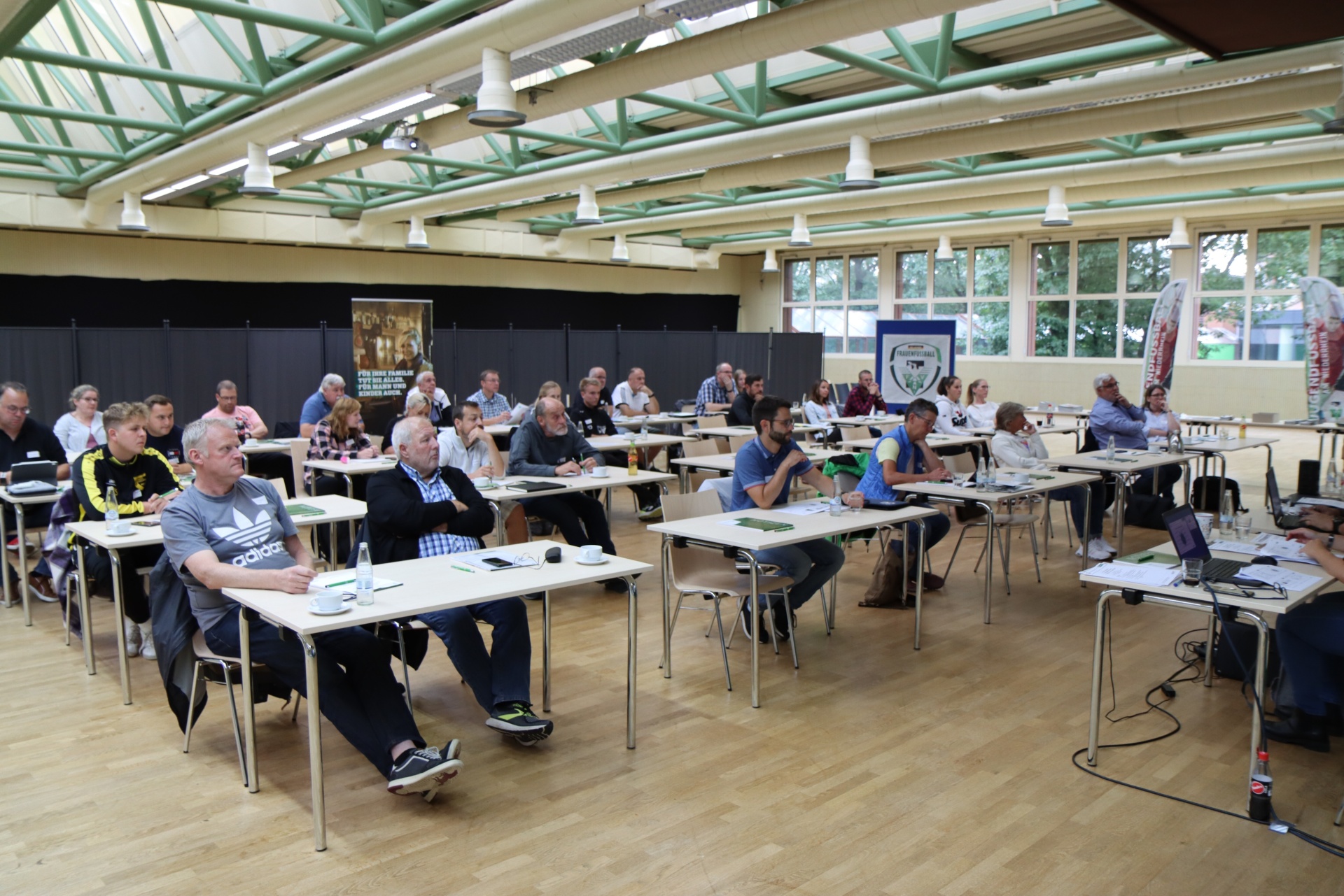 [{"clang_id":1,"value":"Raum mit etwa 30 Personen an Tischen, die an einer Veranstaltung teilnehmen. Im Hintergrund Fenster und eine grüne Stahlkonstruktion."}]