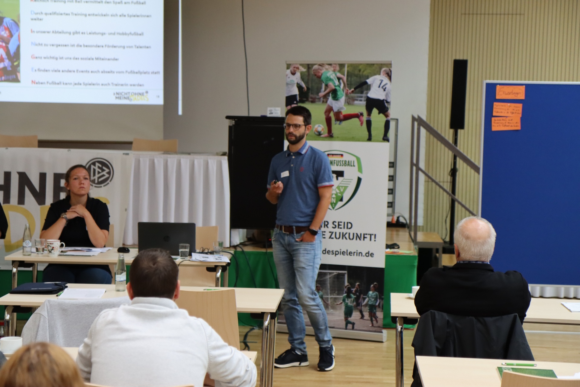 [{"clang_id":1,"value":"Mann hält Vortrag vor Publikum in einem Raum. Im Hintergrund eine Projektion mit Fußballmotiven und Text. Menschen sitzen an Tischen."}]
