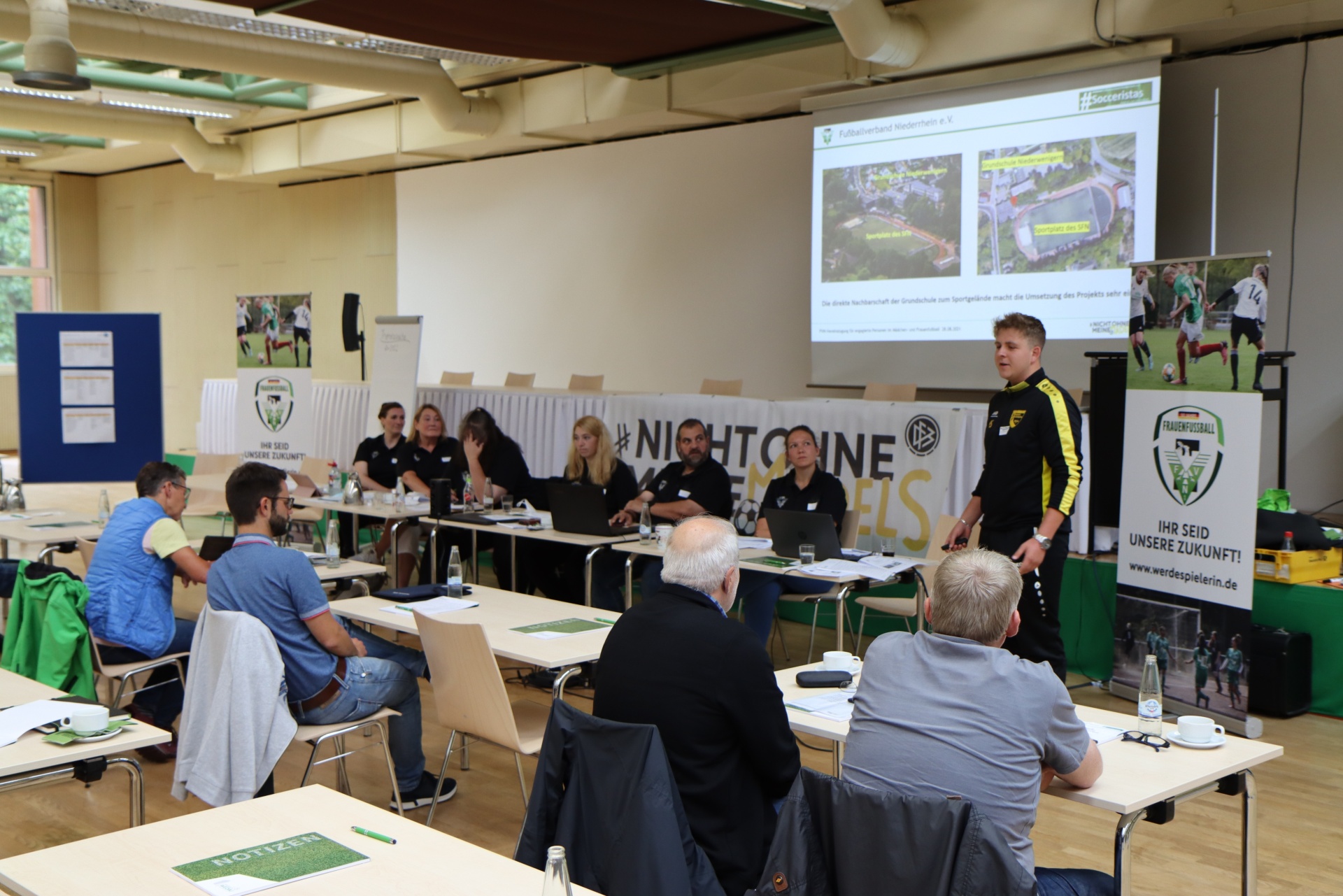 [{"clang_id":1,"value":"Versammlung in Sporthalle. Mann steht und präsentiert Folie mit Planzeichnung. Um ihn herum sitzen ca. 15 Personen an Tischen. Hinter ihm Wand mit Sportlern."}]