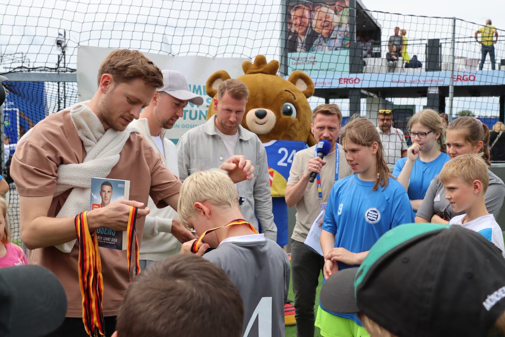 [{"clang_id":1,"value":"Fußballspieler signiert Trikot eines jungen Fans. Im Hintergrund stehen weitere Kinder und Erwachsene, sowie eine Bären-Maskotte."}]