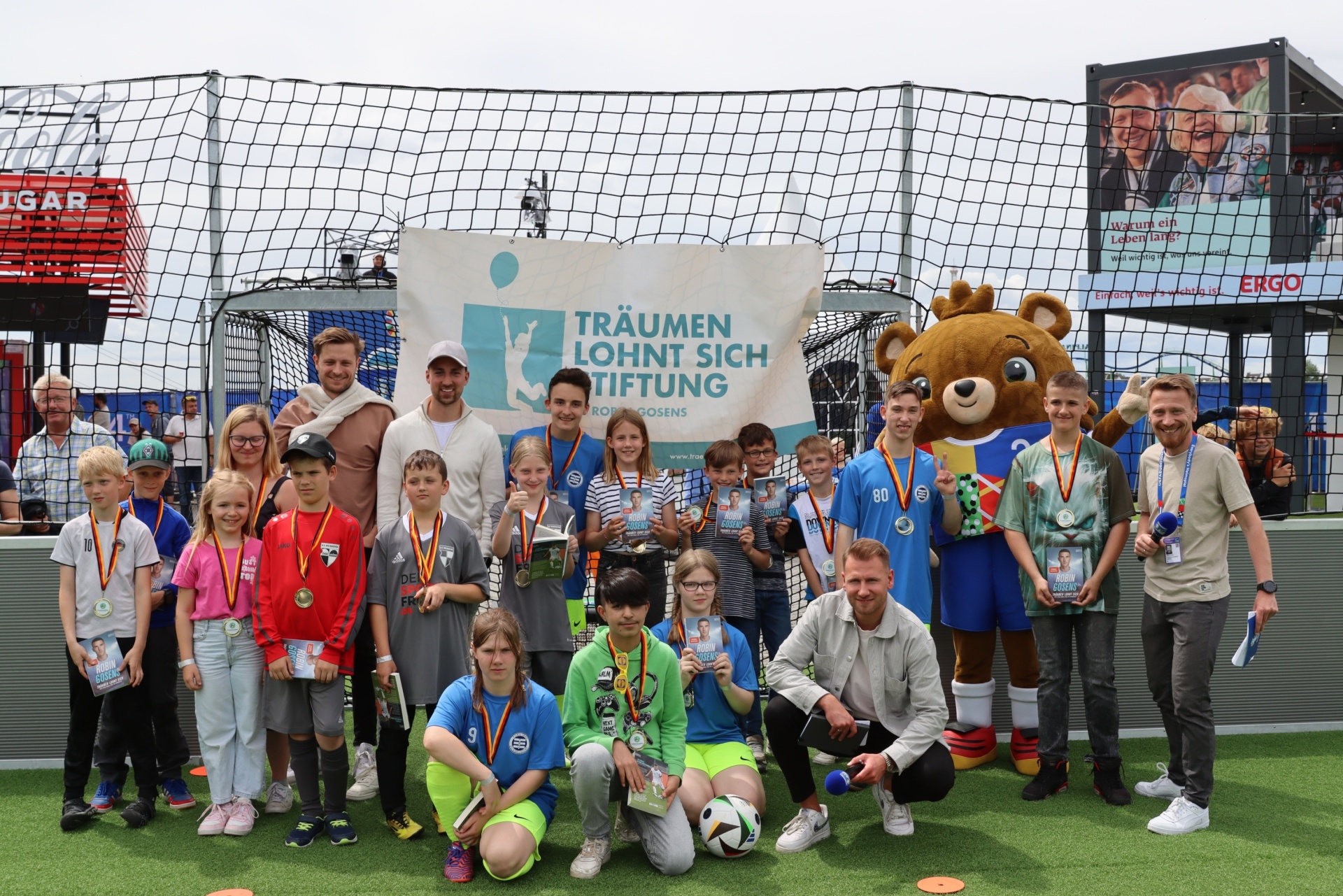 [{"clang_id":1,"value":"Gruppe Kinder mit Medaillen und Betreuer vor Fußballtor mit Banner \"Träumen lohnt sich Stiftung\". Maskottchen-Bär steht mit dabei."}]
