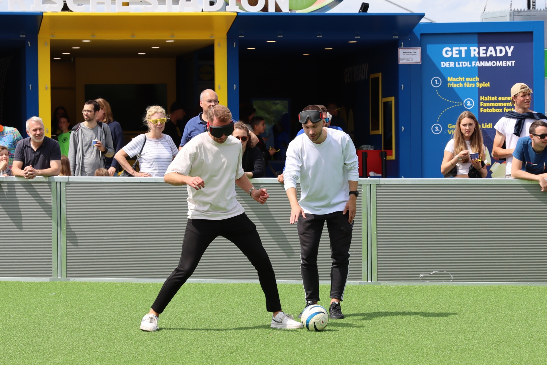 [{"clang_id":1,"value":"Zwei Personen mit Augenbinde und VR-Brille treten auf einem grünen Rasen gegen einen Fußball an. Im Hintergrund eine Zuschauermenge und eine blaue Werbefläche."}]