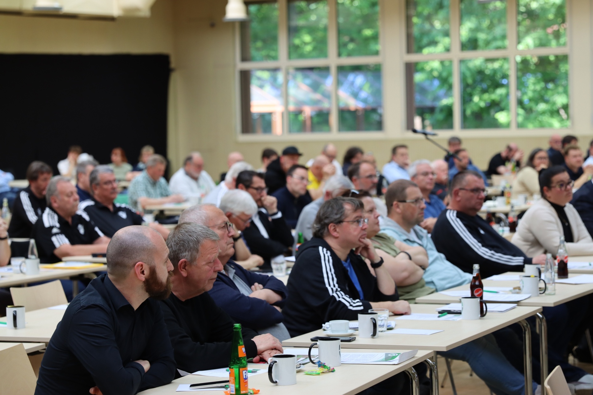 [{"clang_id":1,"value":"Große Menschengruppe sitzt in einem hellen Saal an Tischen. Sie hören einer Präsentation zu. Viele tragen dunkle Kleidung."}]