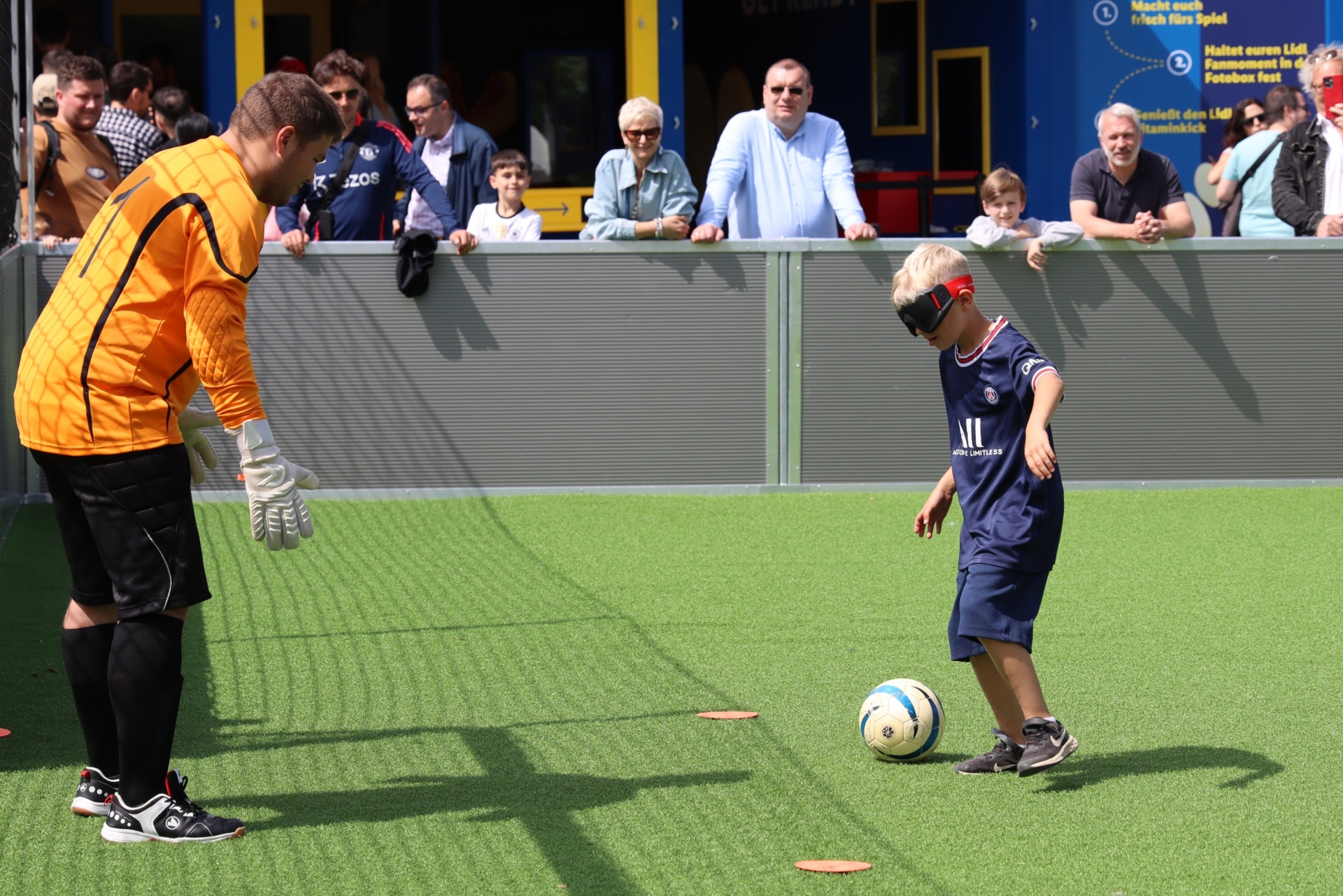[{"clang_id":1,"value":"Junge mit Augenbinde schießt Fußball auf Torwart. Im Hintergrund Zuschauer auf einer Tribüne. Kunstrasenplatz mit Markierungen."}]