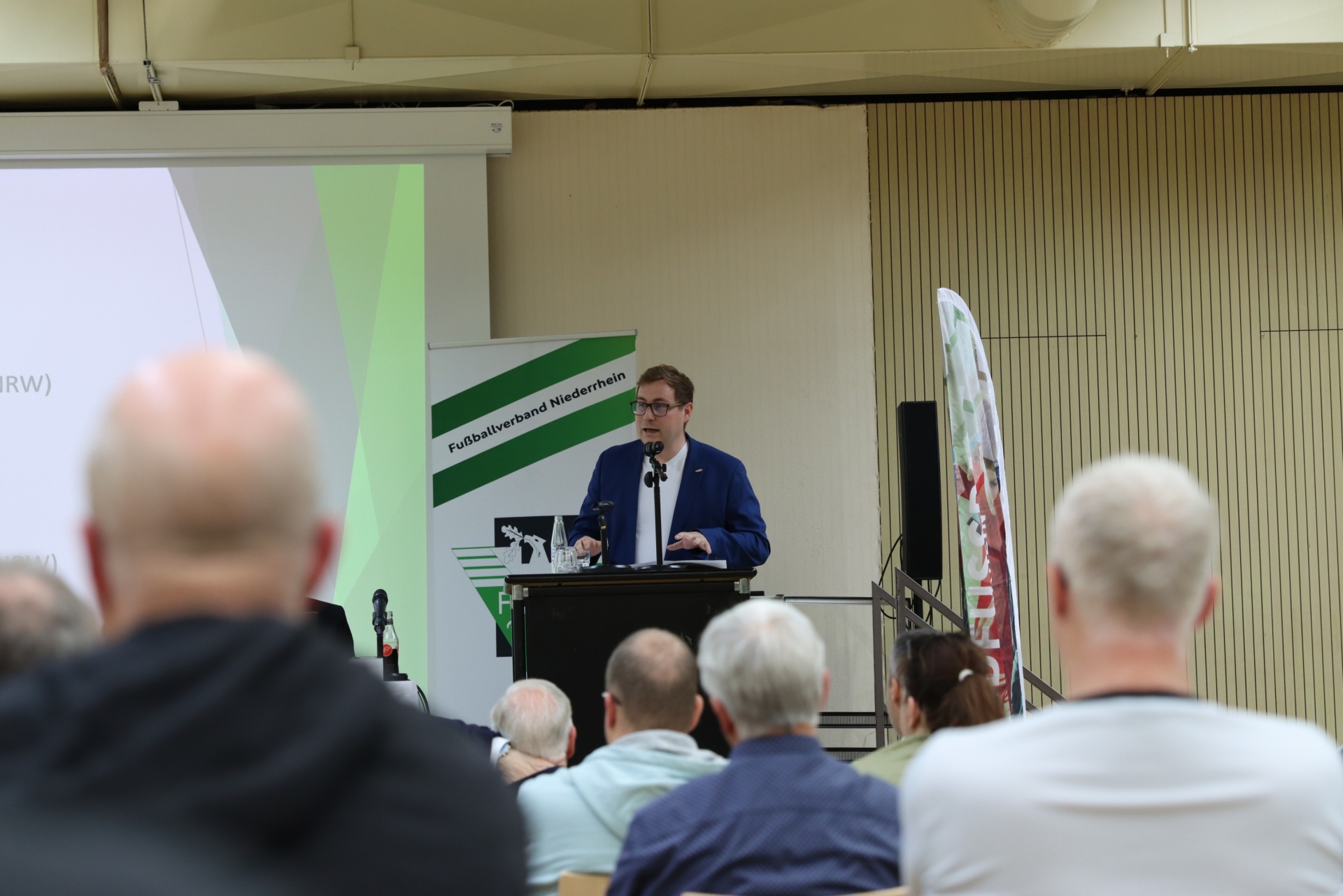 [{"clang_id":1,"value":"Mann am Rednerpult vor Publikum. Im Hintergrund eine Leinwand und Banner mit Schriftzug \"Fußballverband Niederrhein\"."}]