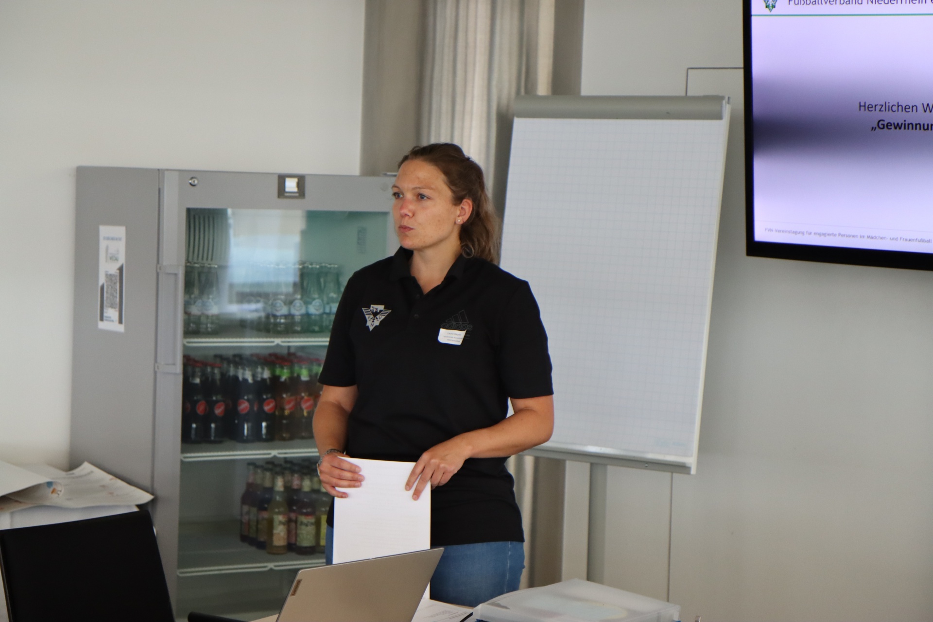[{"clang_id":1,"value":"Frau mit Papieren vor Whiteboard und Kühlschrank mit Getränken. Präsentation im Hintergrund."}]
