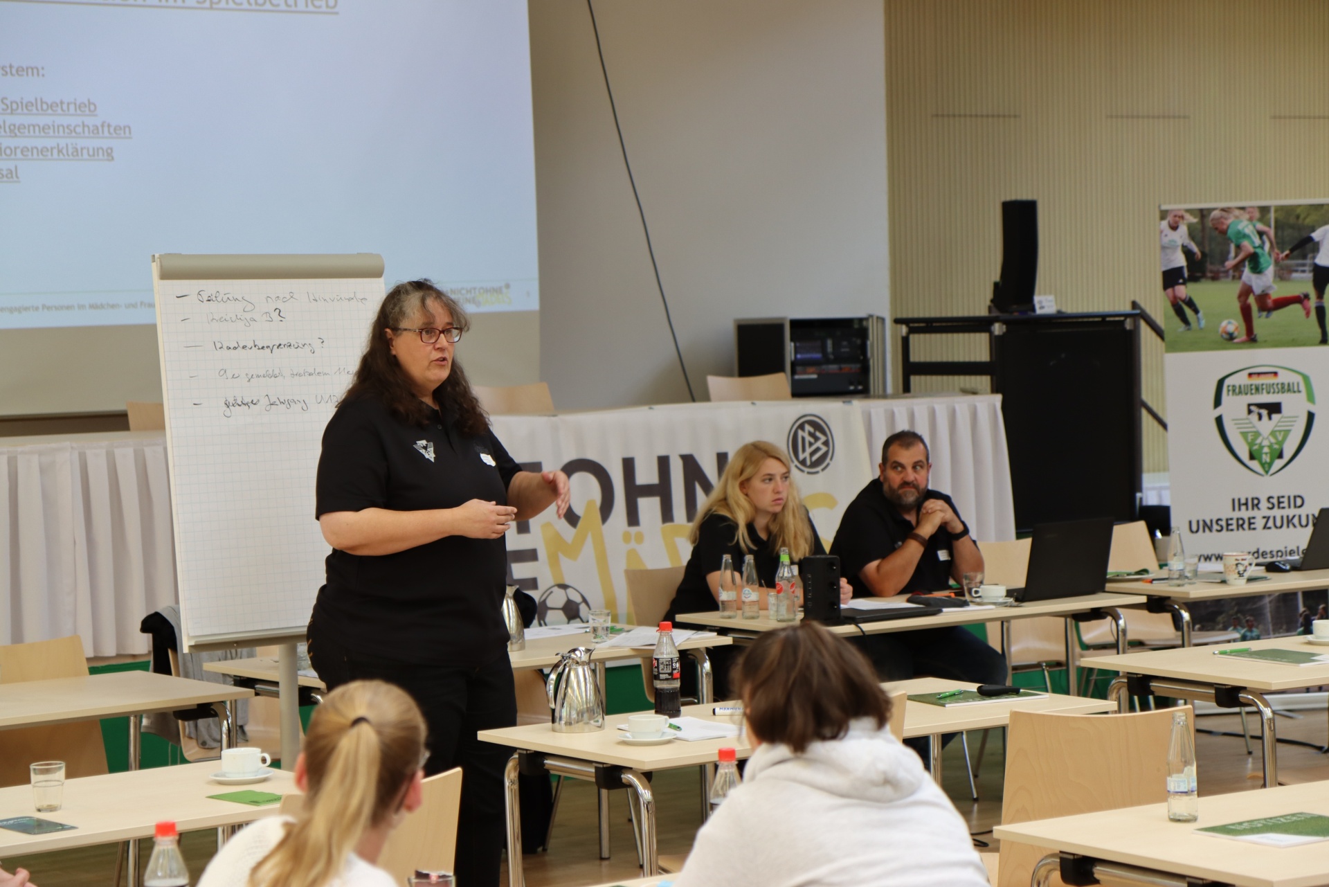 [{"clang_id":1,"value":"Frau hält Vortrag vor Gruppe in Klassenzimmer. Präsentationsfolie im Hintergrund, Tische mit Teilnehmern, Wasserflaschen und Schreibmaterialien."}]
