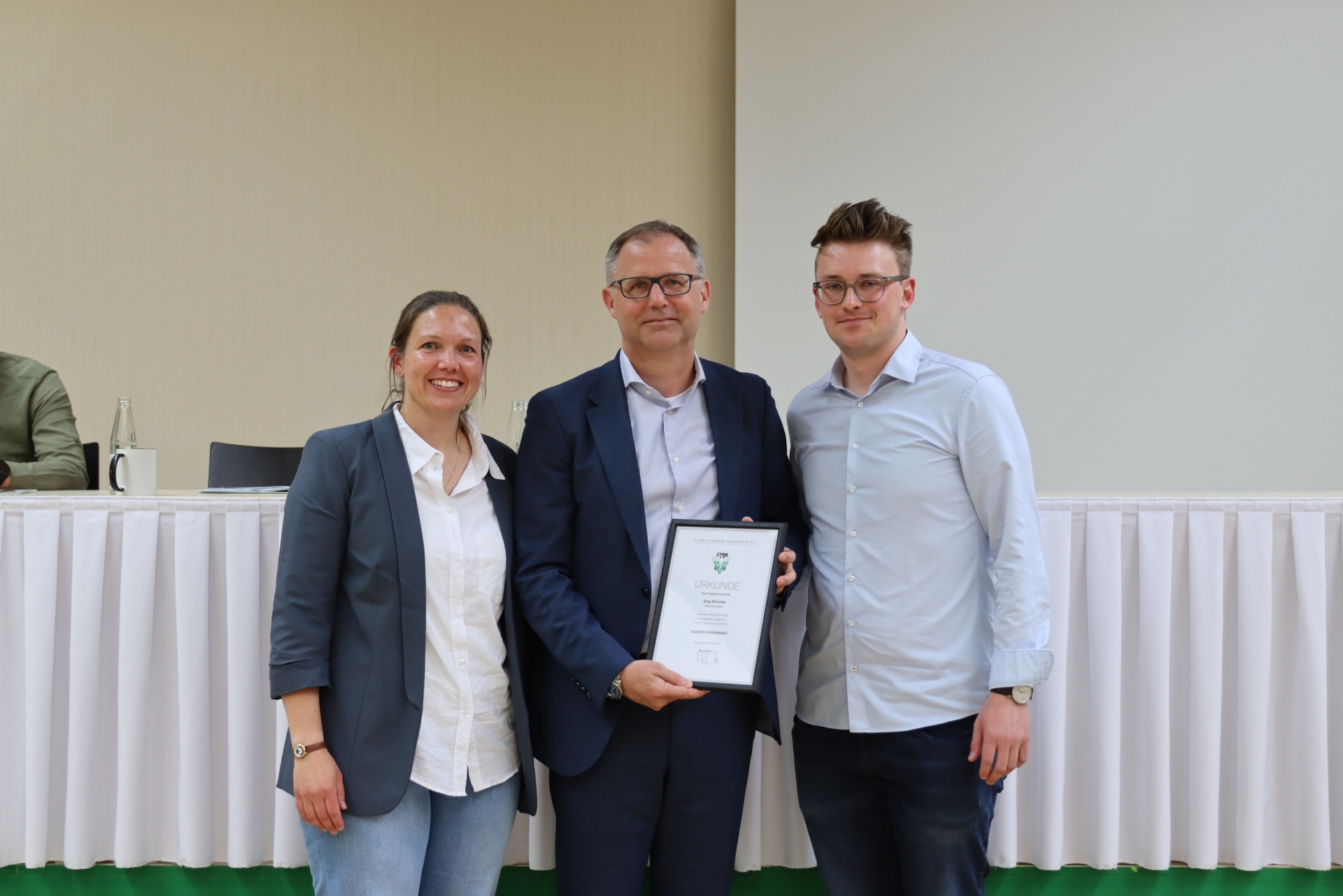 [{"clang_id":1,"value":"Drei Personen stehen nebeneinander vor einem Tisch mit weißer Tischdecke. Die Frau links trägt Blazer, Jeans und ein Hemd. In der Mitte hält ein Mann in Anzug und Hemd eine Urkunde. Rechts steht ein Mann mit Hemd und Jeans."}]