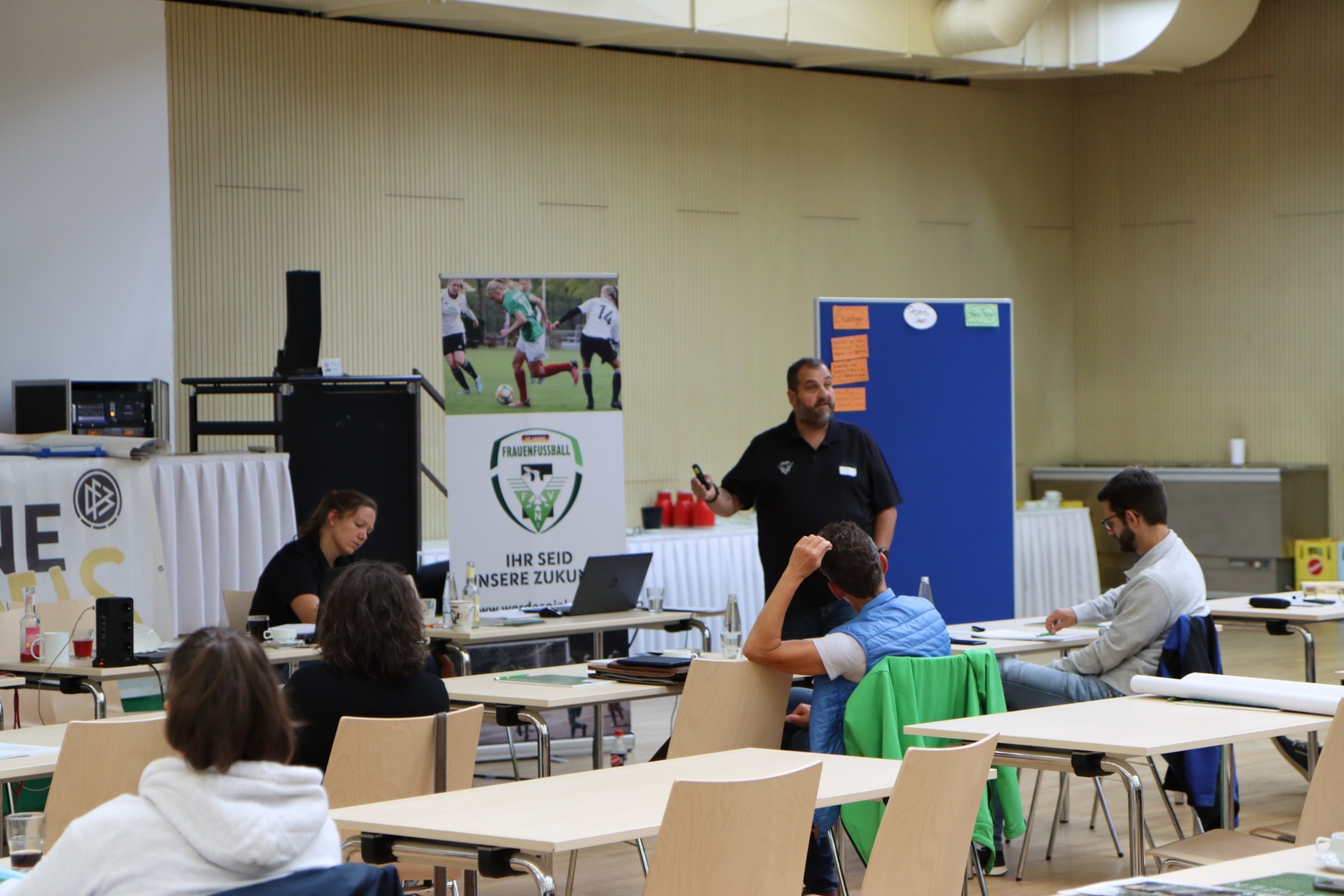 [{"clang_id":1,"value":"Gymnastikraum mit Seminar: Mann spricht vor kleinen Gruppen an Tischen. Im Hintergrund Banner mit Fußballmotiv und Text."}]