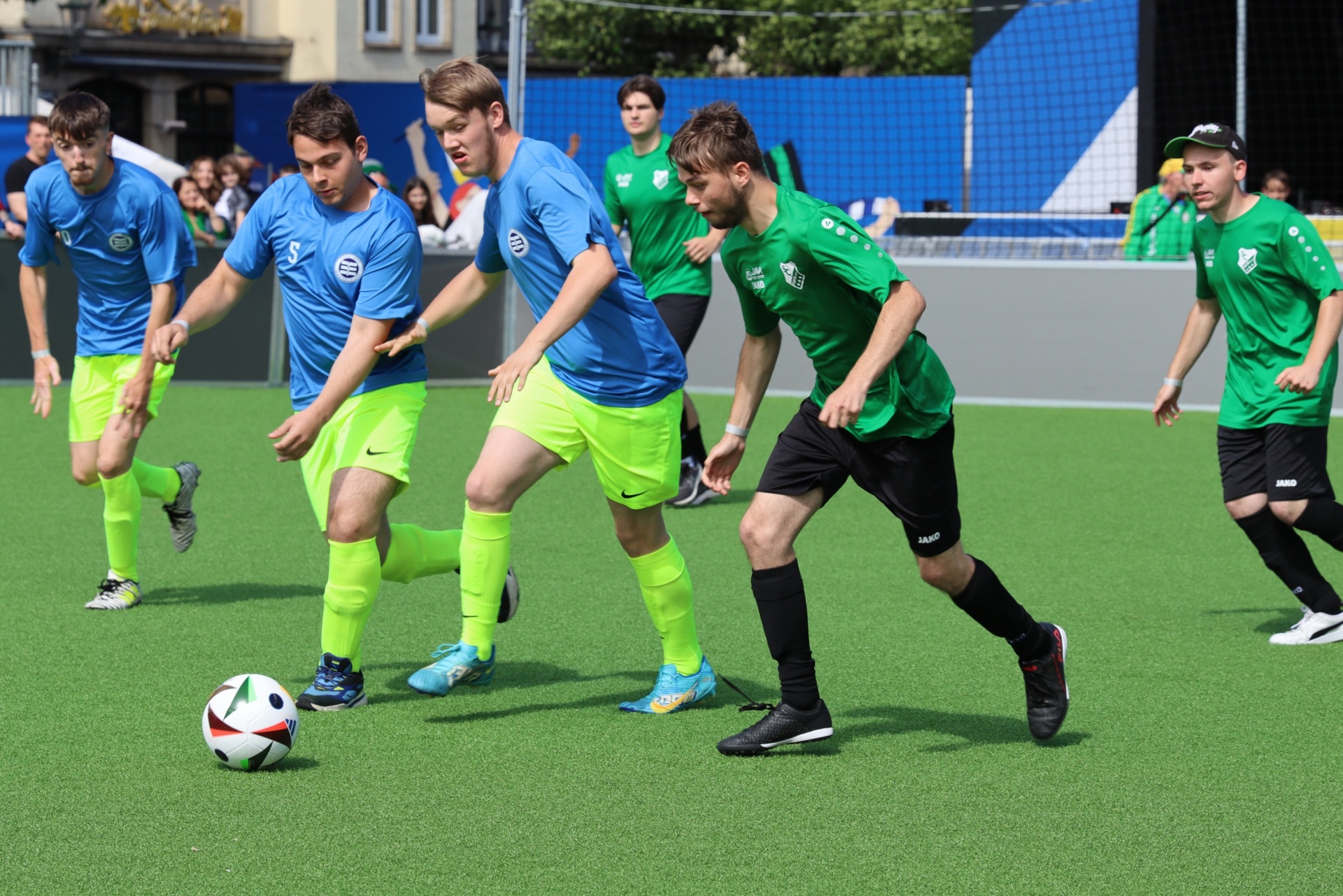 [{"clang_id":1,"value":"Fußballspiel auf grünem Kunstrasen. Zwei Mannschaften in blau und grün kämpfen um den Ball. Mehrere Spieler in Aktion. Zuschauer im Hintergrund."}]