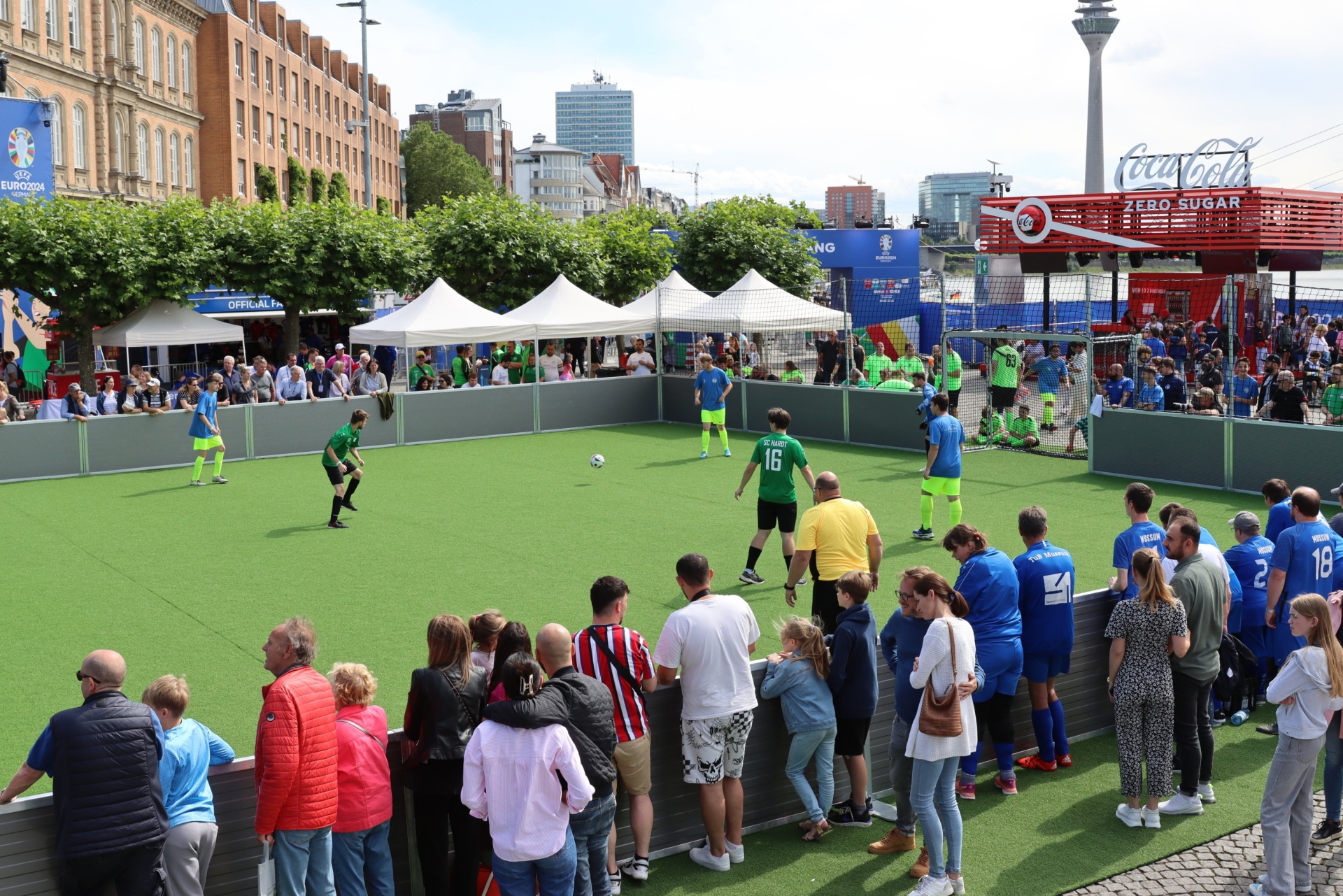 [{"clang_id":1,"value":"Fußballspiel auf Kunstrasenfeld mit Zuschauern. Im Hintergrund Gebäude, Bäume und ein Sendemast mit Coca-Cola Werbung."}]