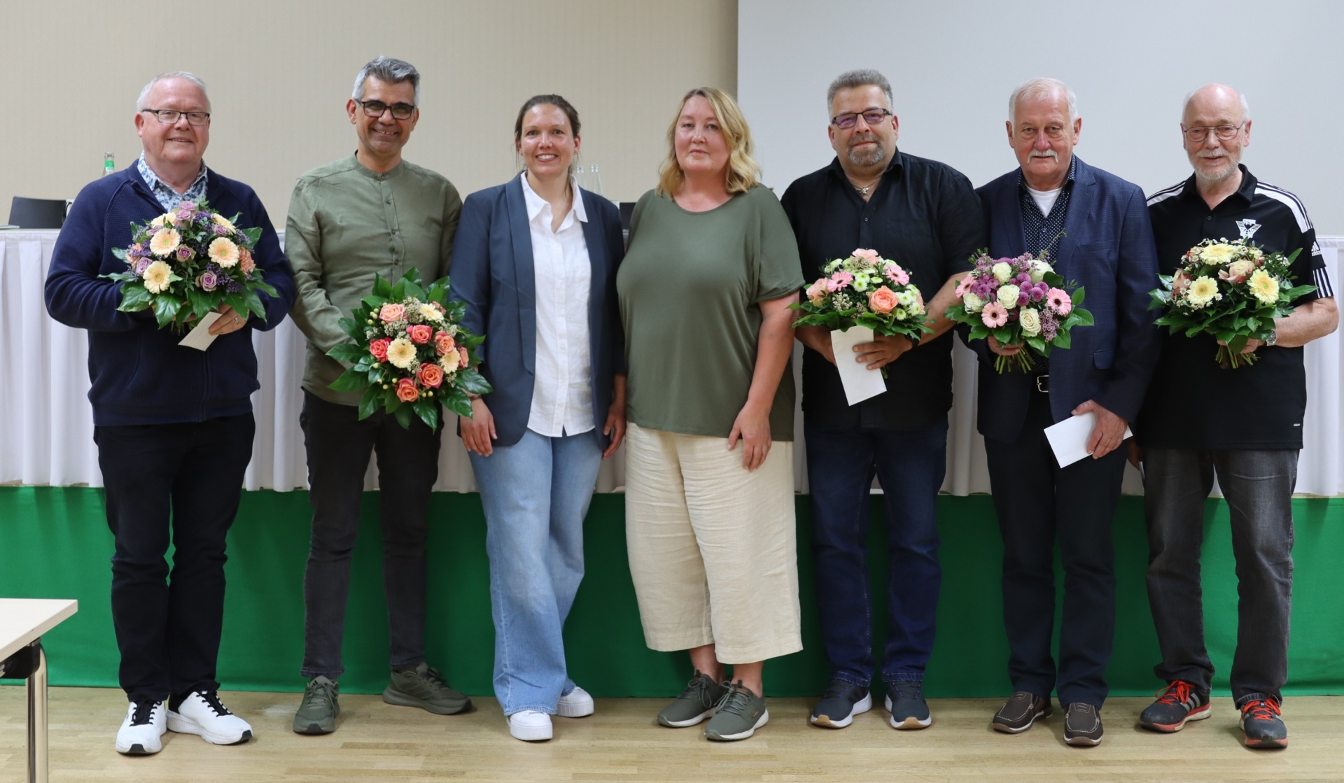 [{"clang_id":1,"value":"Sieben Personen stehen auf einer Bühne, jeder hält einen Blumenstrauß. Alle tragen Freizeitkleidung. Im Hintergrund eine grüne Wand und eine weiße Leinwand."}]