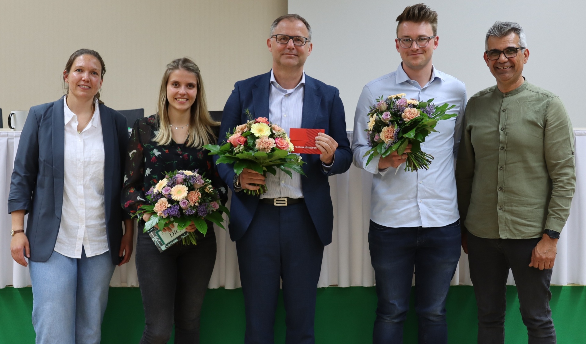 [{"clang_id":1,"value":"Fünf Personen stehen nebeneinander auf einer Bühne. Sie halten Blumensträuße und einen roten Umschlag. Alle tragen formelle Kleidung."}]