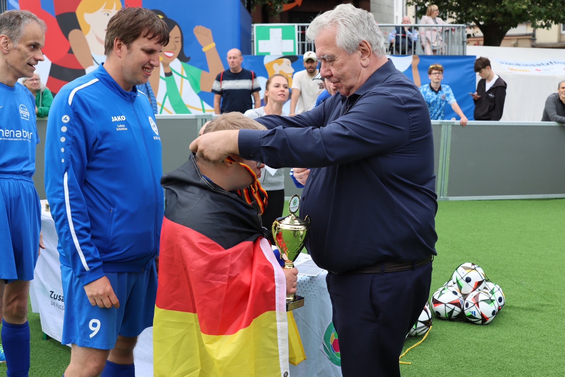 [{"clang_id":1,"value":"Drei Männer stehen auf einem Rasenplatz. Ein Mann in blauem Sportanzug erhält von einem älteren Mann in dunklem Hemd eine Trophäe. Im Hintergrund eine Frau mit Deutschlandflagge."}]