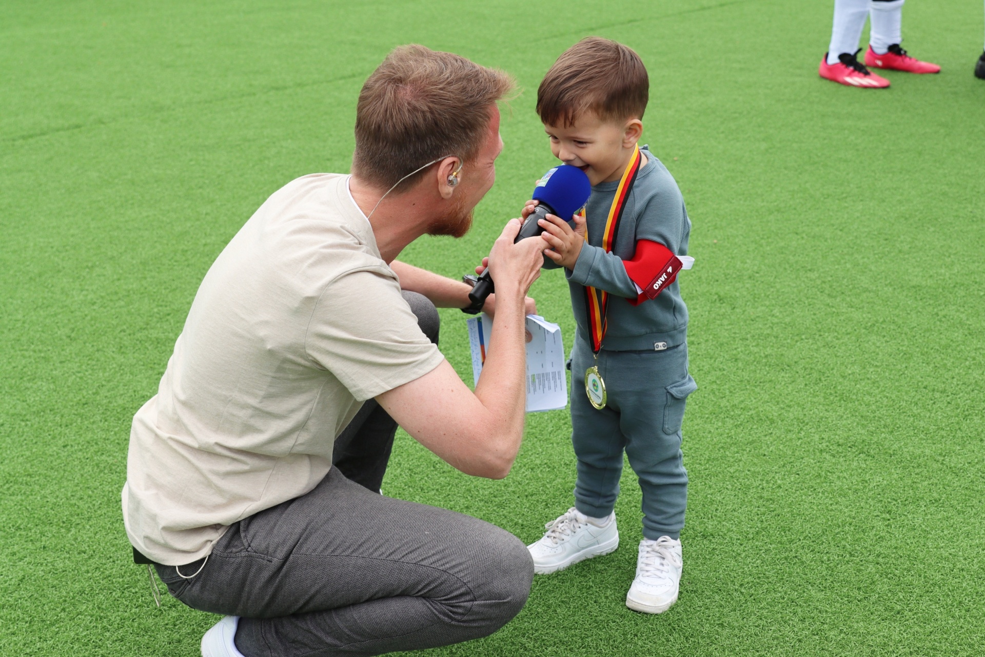 [{"clang_id":1,"value":"Journalist kniet vor einem kleinen Jungen auf grünem Rasen. Er hält ein Mikrofon an den Jungen, der ein Medaillenband trägt."}]