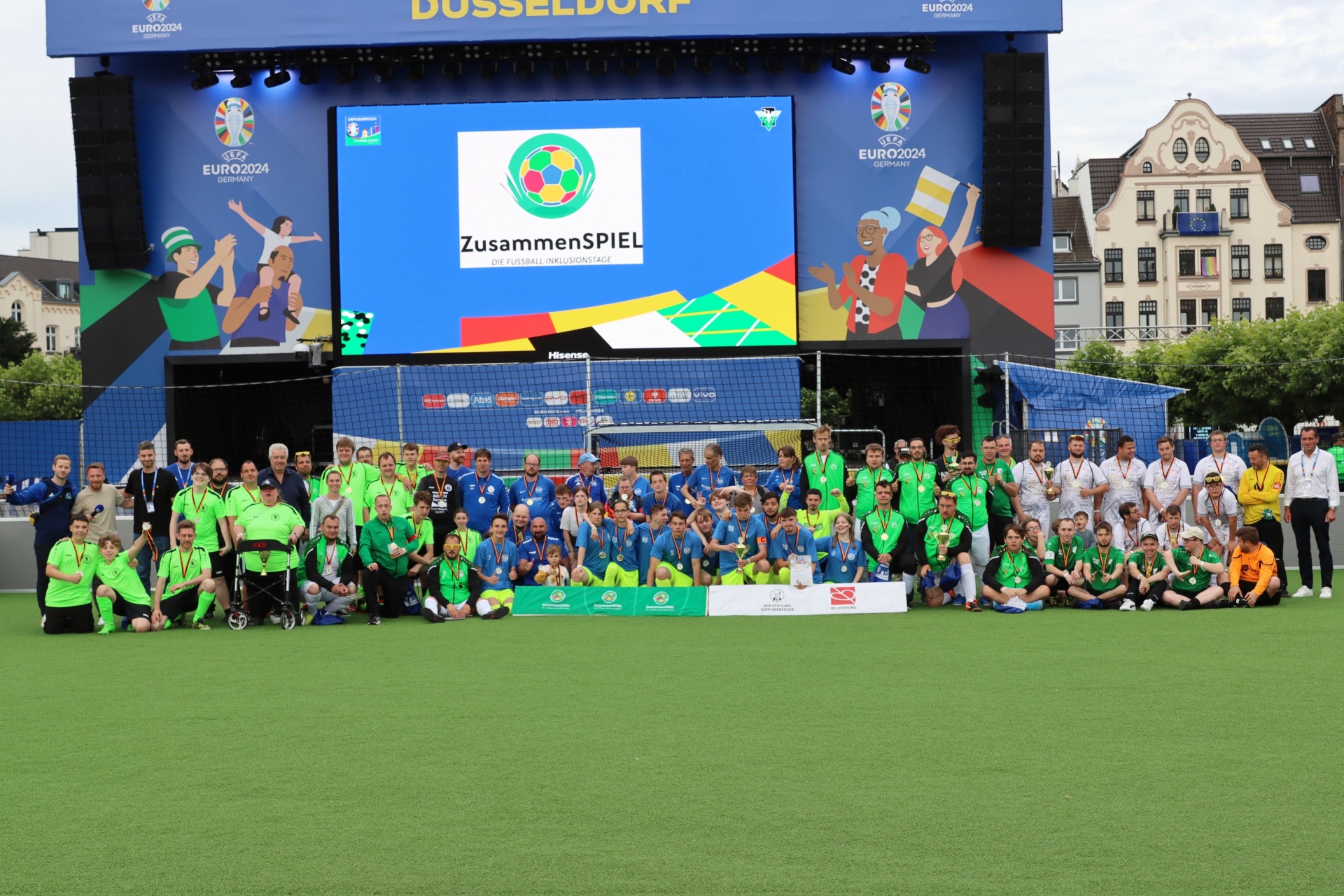 [{"clang_id":1,"value":"Gruppe Menschen vor Großbildschirm mit EURO 2024 Logo und Schriftzug \"ZusammenSPIEL\" auf Fußballfeld in Düsseldorf."}]