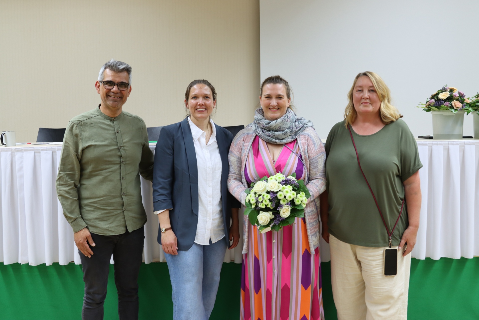 [{"clang_id":1,"value":"Vier Personen stehen nebeneinander vor einem Tisch mit weißer Tischdecke und grünem Stoff. Von links: Mann mit Hemd, Frau mit Blazer, Frau mit Blumenstrauß und Frau mit T-Shirt."}]