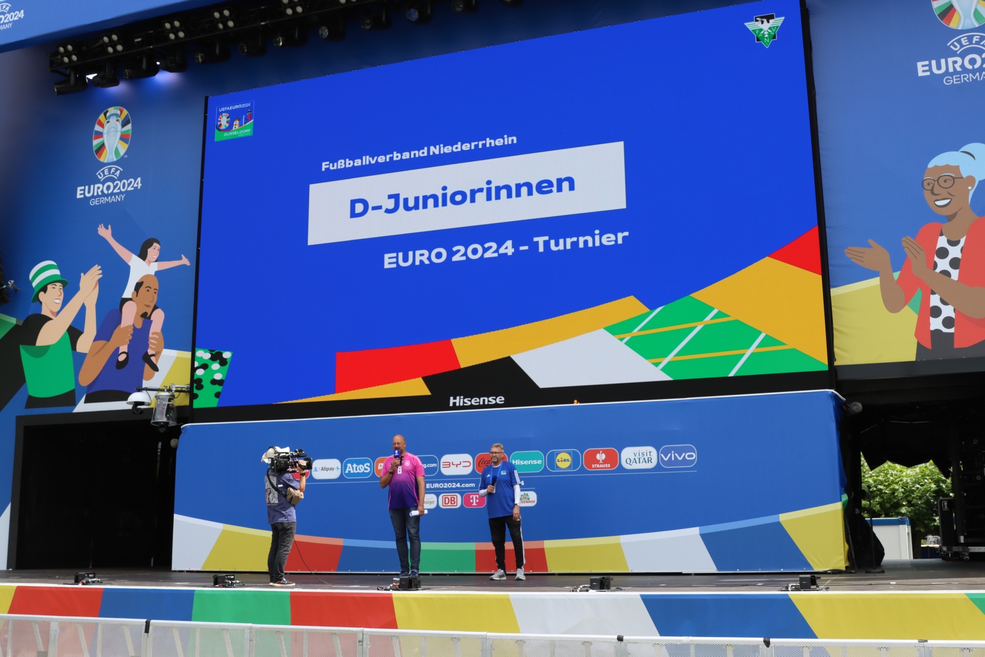 [{"clang_id":1,"value":"Große LED-Wand zeigt \"D-Juniorinnen EURO 2024\" mit Logos und Grafik. Darunter stehen drei Personen auf einer Bühne vor einem Werbebanner."}]