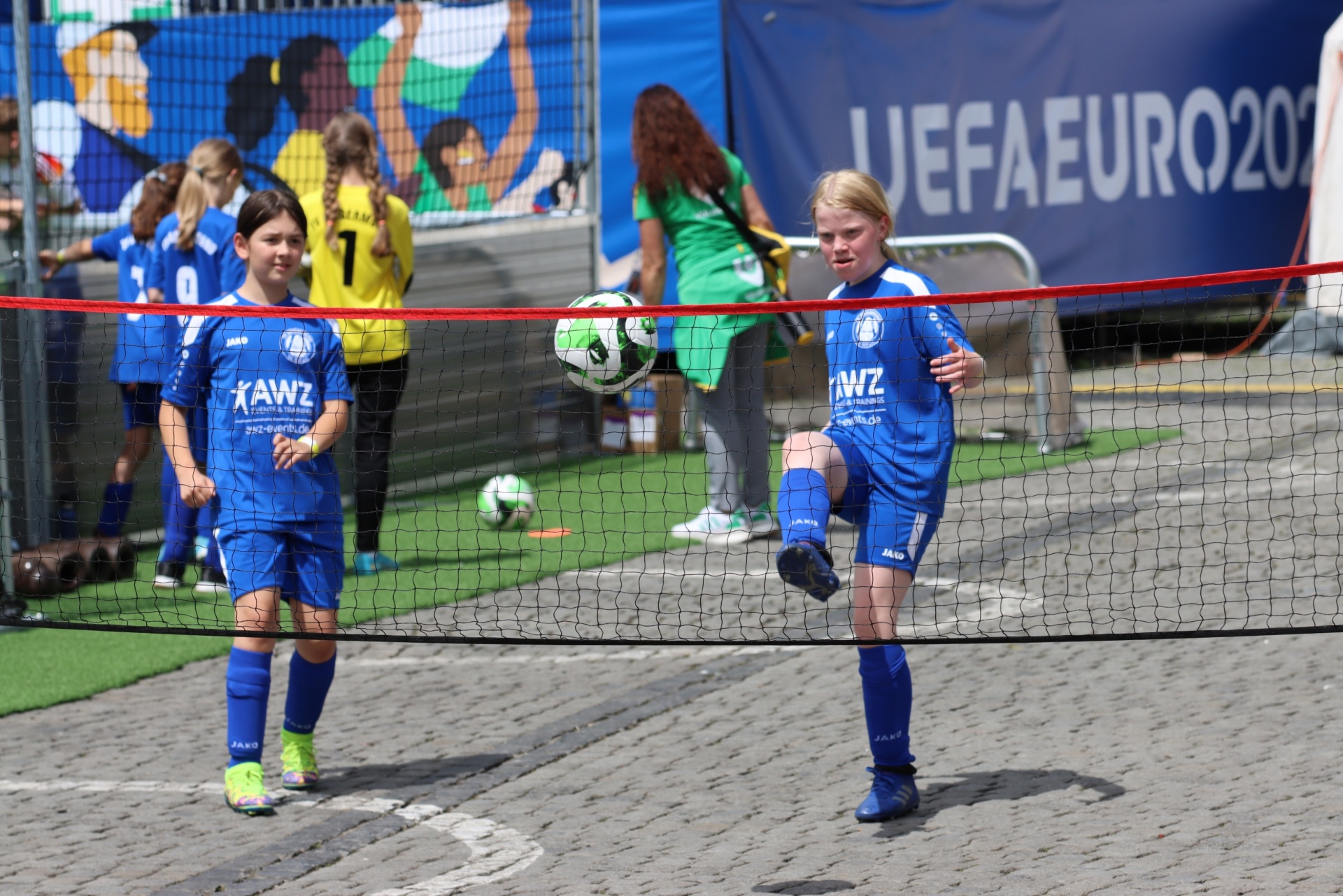 [{"clang_id":1,"value":"Zwei Kinder in blauen Fußballtrikots spielen auf einem gepflasterten Platz. Im Hintergrund eine Werbeplane mit \"UEFA EURO 2020\" und weitere Personen."}]