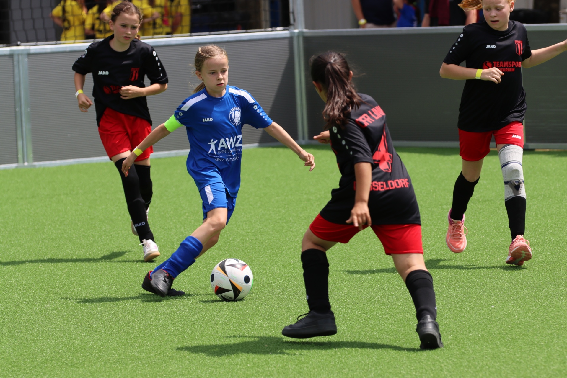 [{"clang_id":1,"value":"Fußballspiel mit vier jungen Spielerinnen auf grünem Kunstrasen. Eine Spielerin in blau dribbelt den Ball, eine in rot steht ihr gegenüber, die anderen sind im Hintergrund."}]