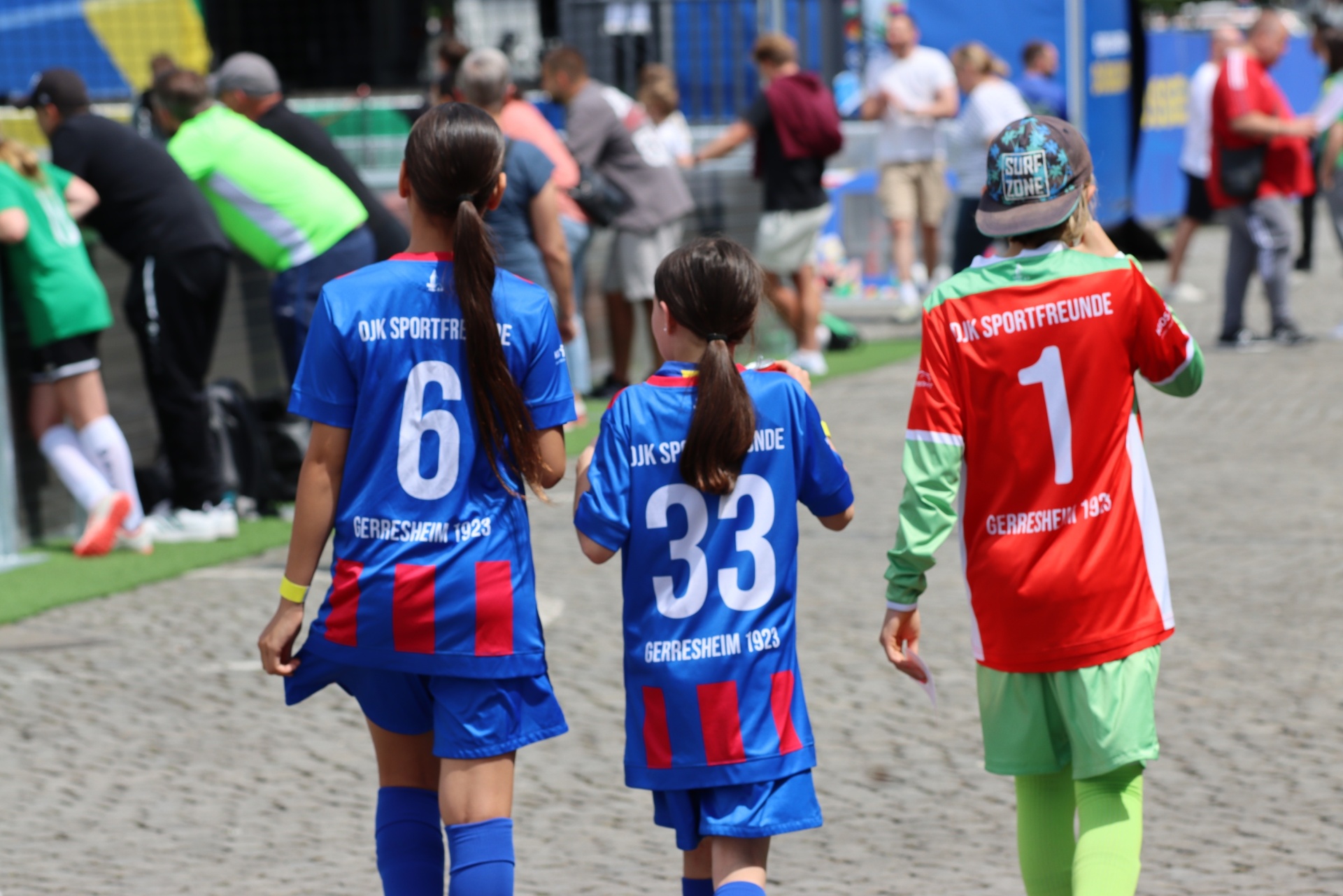 [{"clang_id":1,"value":"Drei junge Fußballspielerinnen mit Trikots der Nummern 6, 33 und 1 gehen auf einem Rasenplatz, umgeben von Zuschauern und Begleitpersonal."}]