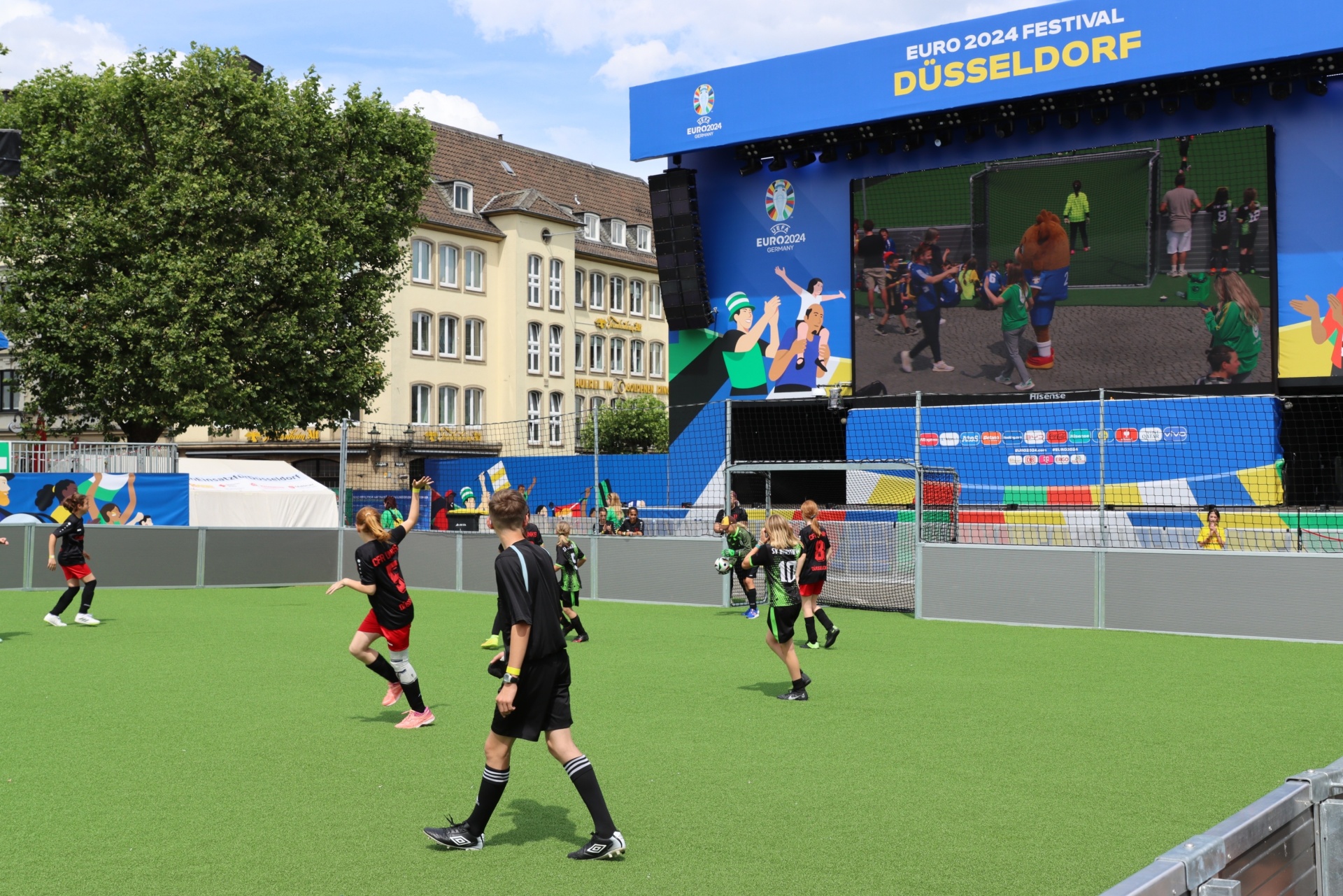 [{"clang_id":1,"value":"Fußballspiel auf Kunstrasen vor einer großen Leinwand mit dem Logo \"EURO 2024 Festival Düsseldorf\". Im Hintergrund Gebäude und Bäume."}]