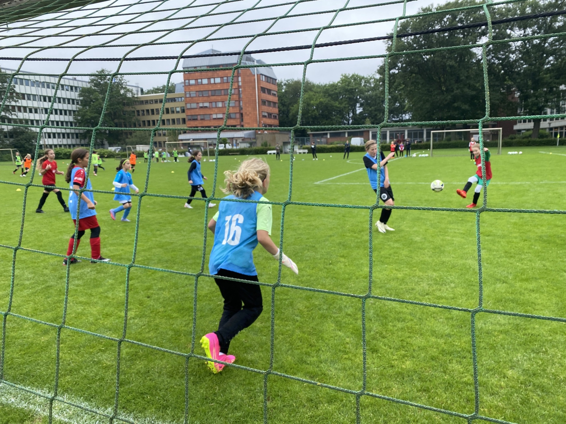 [{"clang_id":1,"value":"Fußballspiel mit Kindern auf grünem Rasenfeld. Im Vordergrund ein Tor mit Netz. Ein Mädchen in blauem Trikot mit Nummer 16 steht davor. Im Hintergrund Gebäude und weitere Spieler."}]