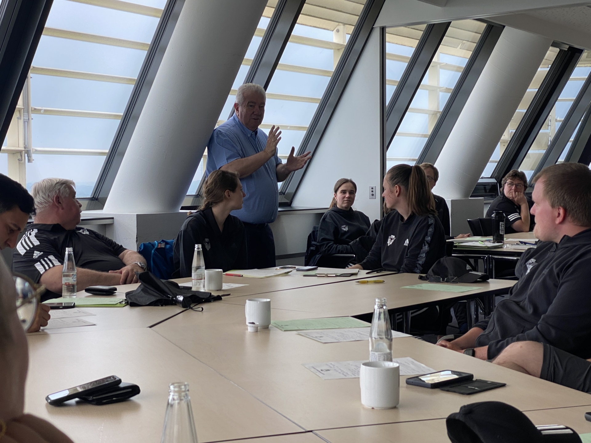 [{"clang_id":1,"value":"Besprechung mit Sportlern: Eine Gruppe, meist in dunkler Sportkleidung, sitzt um einen Konferenztisch. Ein Mann steht vor der Gruppe und spricht. Große Fenster im Hintergrund."}]