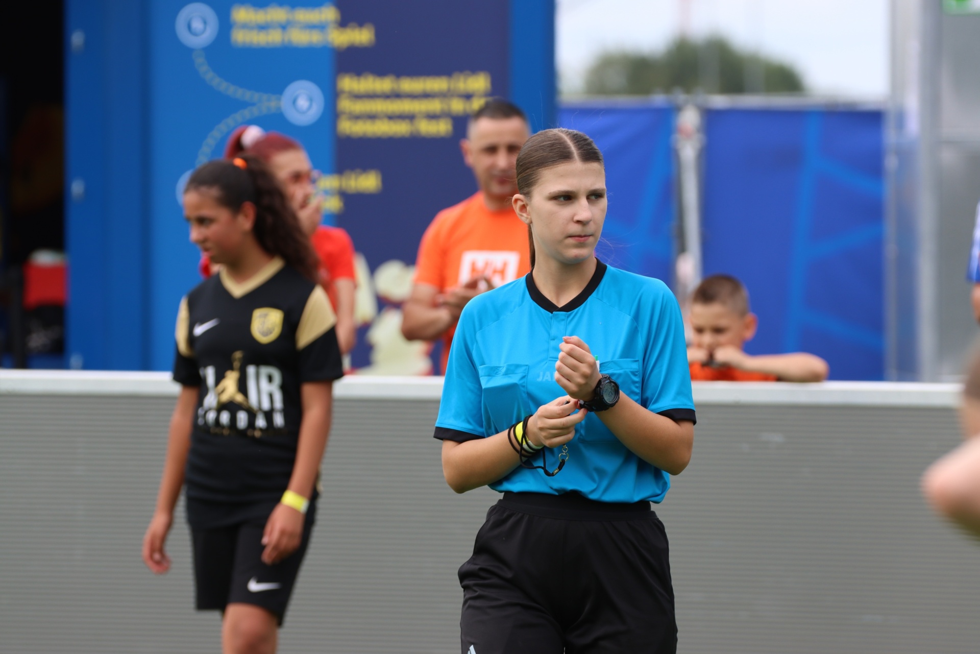 [{"clang_id":1,"value":"Fußballspiel: Schiedsrichterin mit Pfeife und Uhr am Handgelenk steht vor einer Bande. Im Hintergrund Spieler und Zuschauer."}]